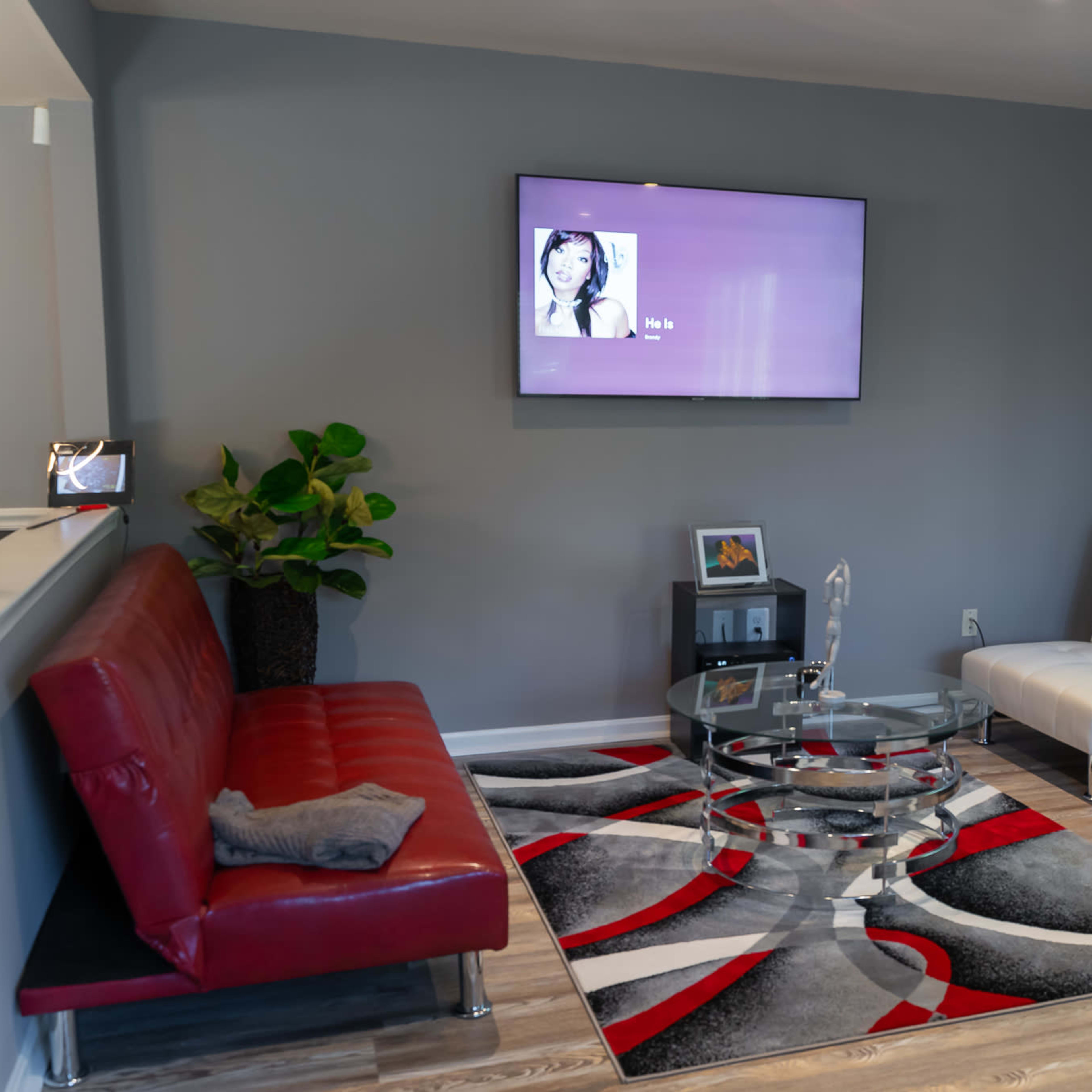 A modern living room featuring a red sofa, a black and white sofa, a glass coffee table on a patterned rug, and a wall-mounted television.