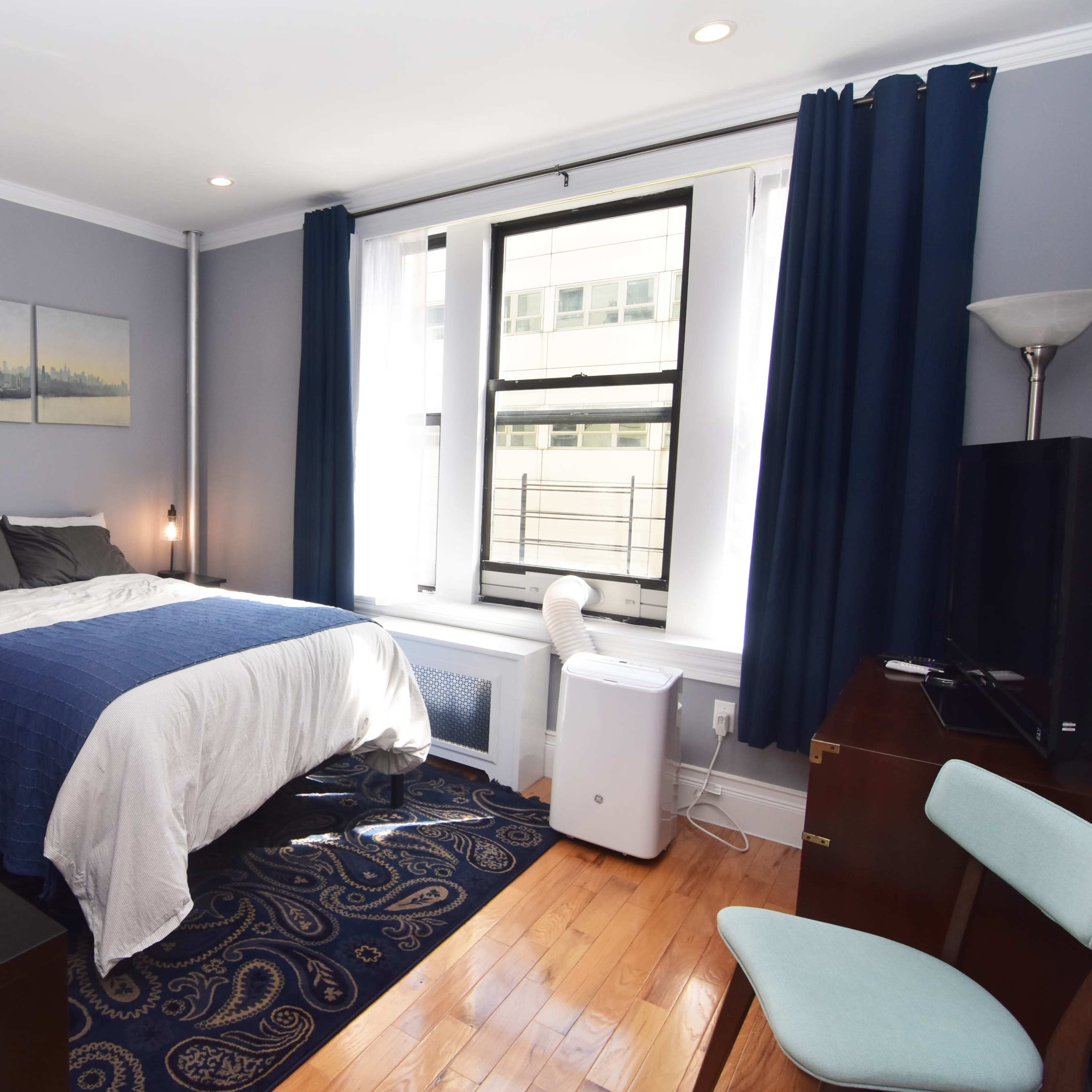 A neatly arranged bedroom featuring a bed, desk, plant, and window with curtains.