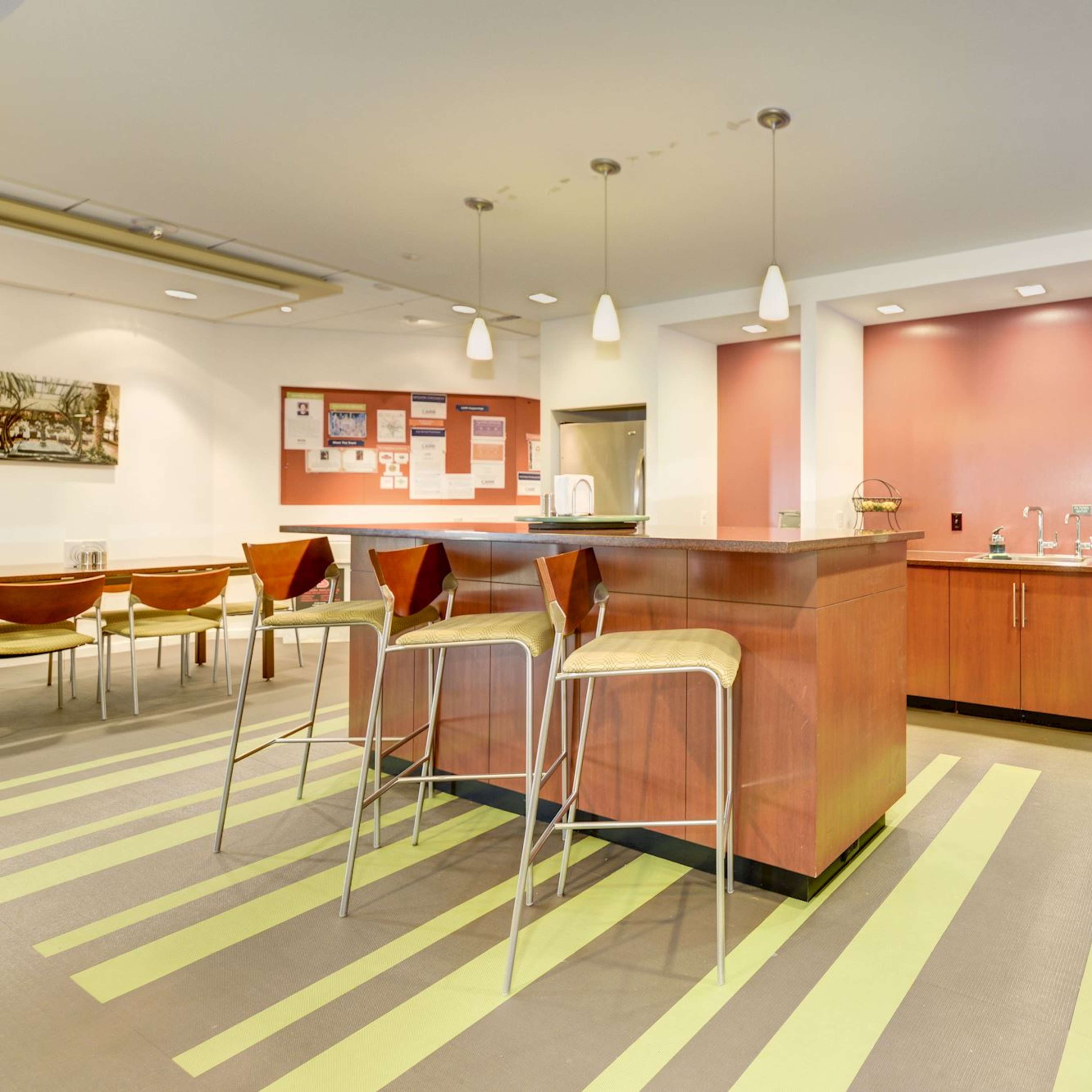 A modern kitchen area features a central island with bar stools, cabinetry, and appliances, with a patterned floor and wall-mounted bulletin board.