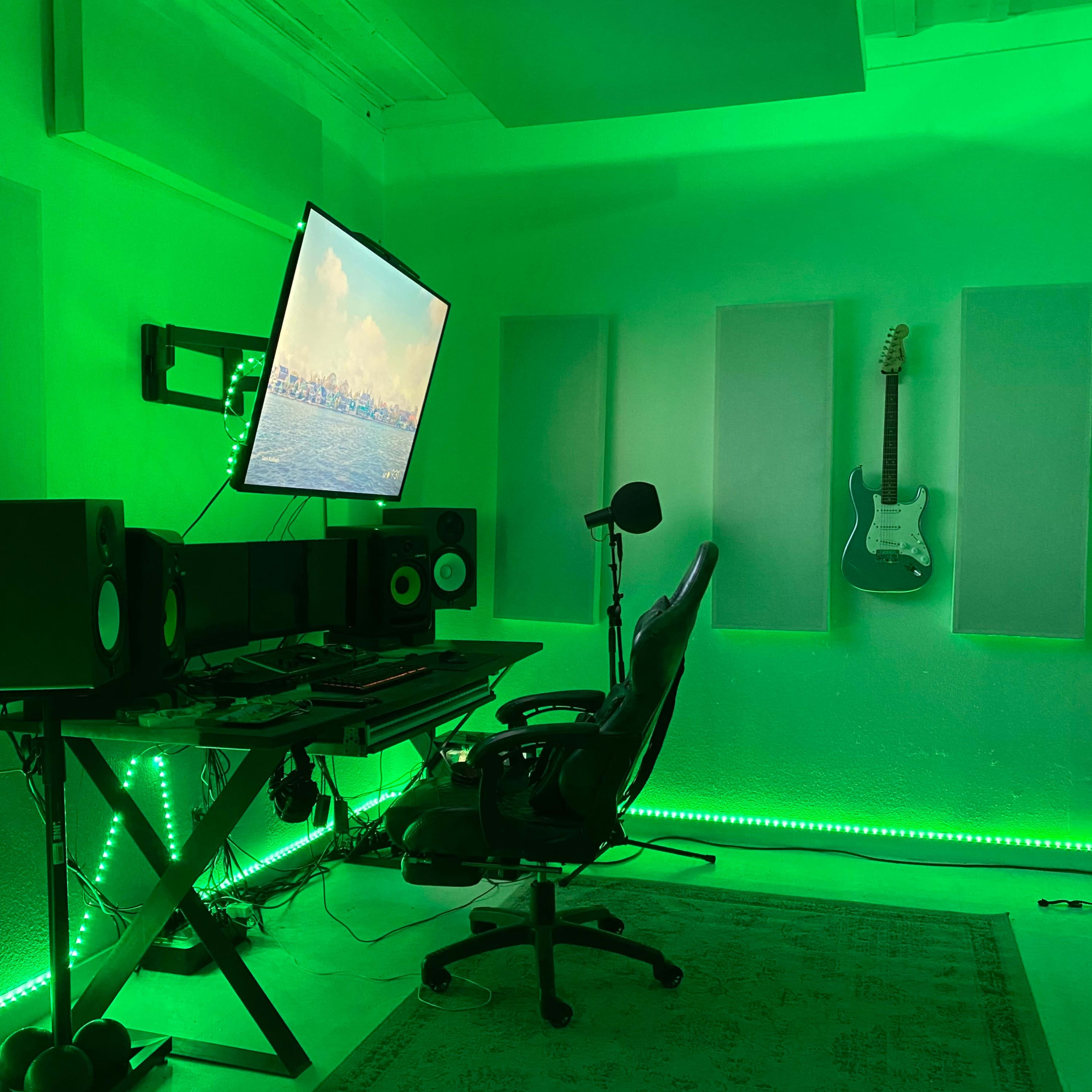 A music production studio with a green color scheme, featuring a desk with audio equipment, a swivel chair, a large monitor displaying a landscape, and an electric guitar mounted on the wall.