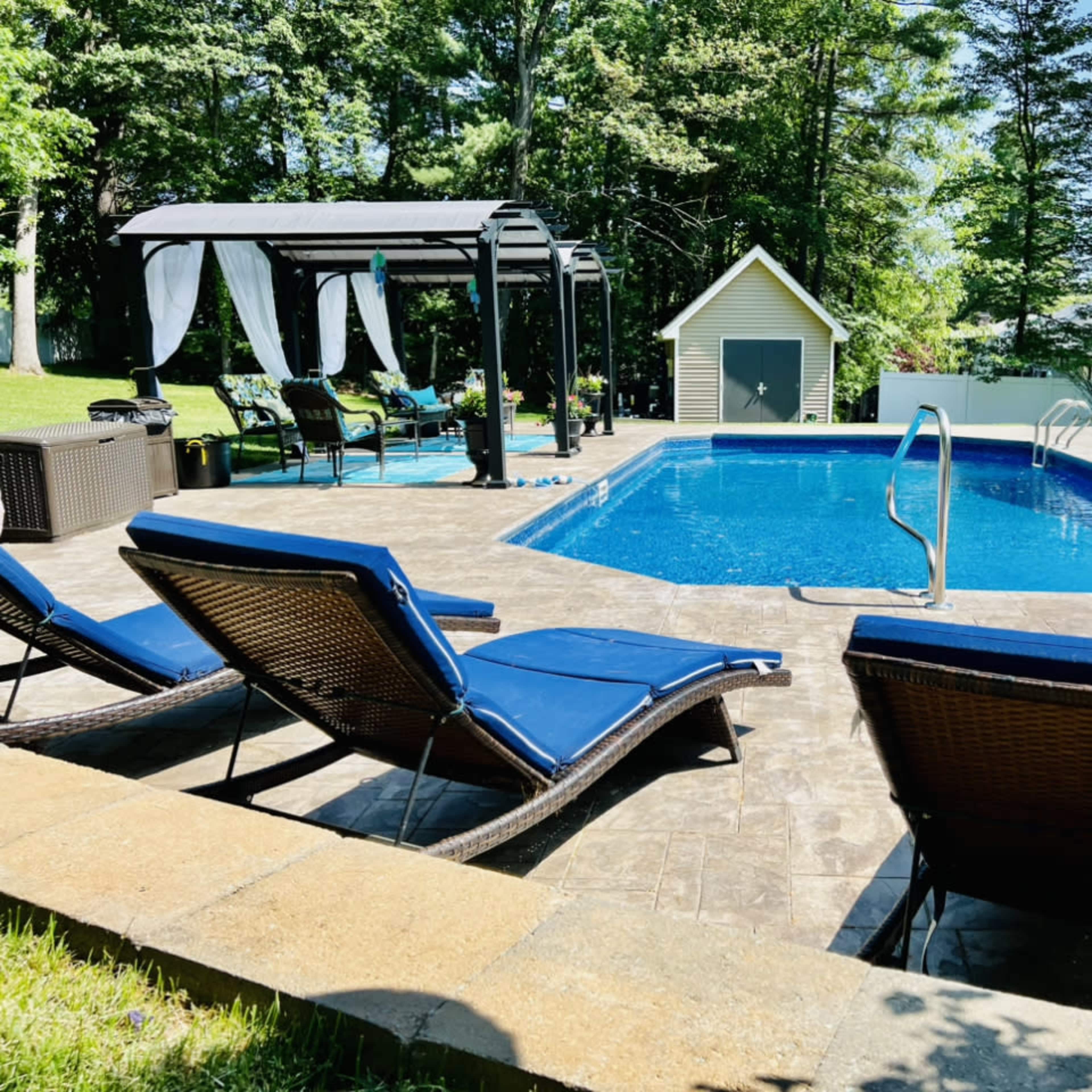 A backyard swimming pool surrounded by lounge chairs, a gazebo, and a shed, with trees in the background.