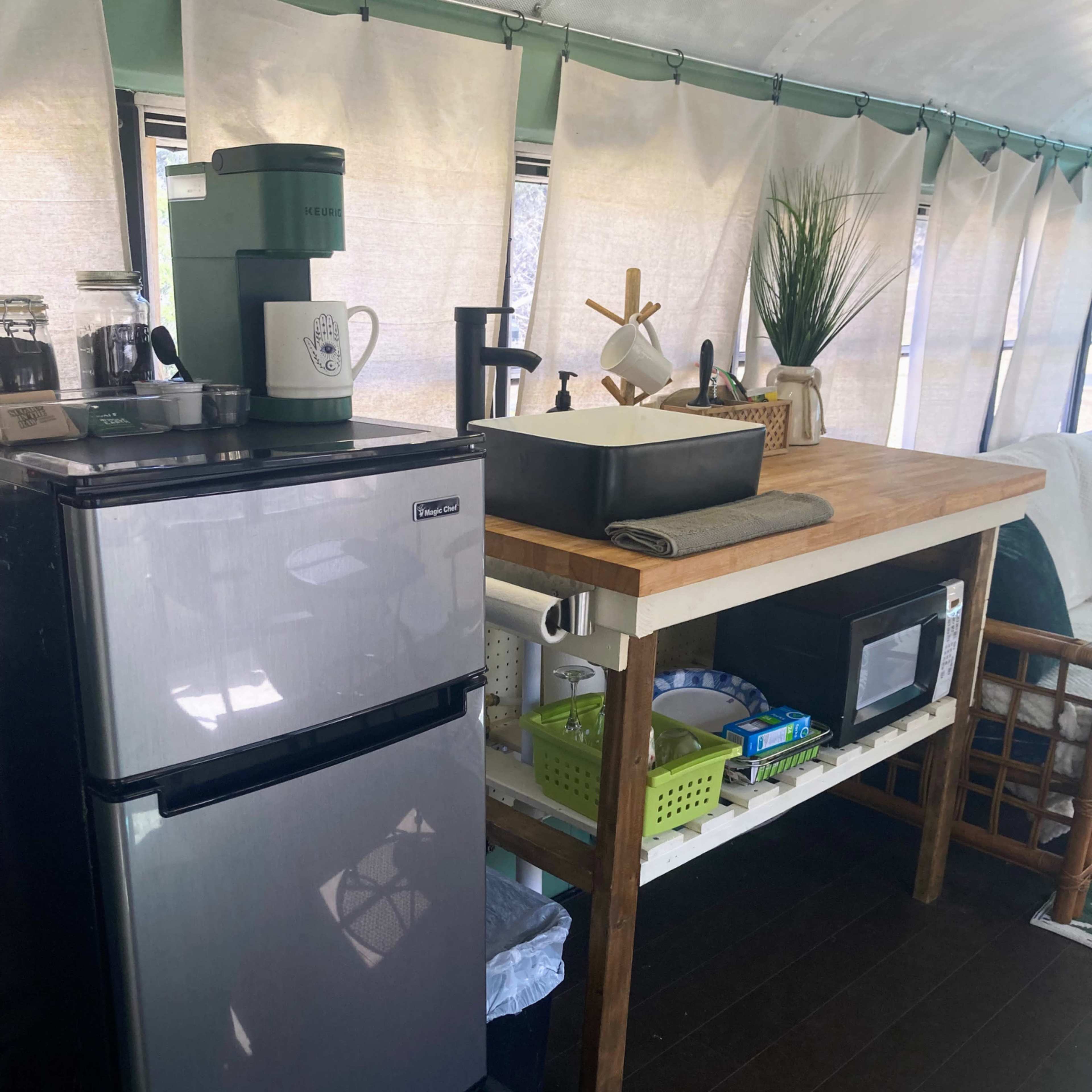 A small kitchen area inside a converted bus, featuring a refrigerator, a wooden table with a sink, and various kitchen items organized on shelves.