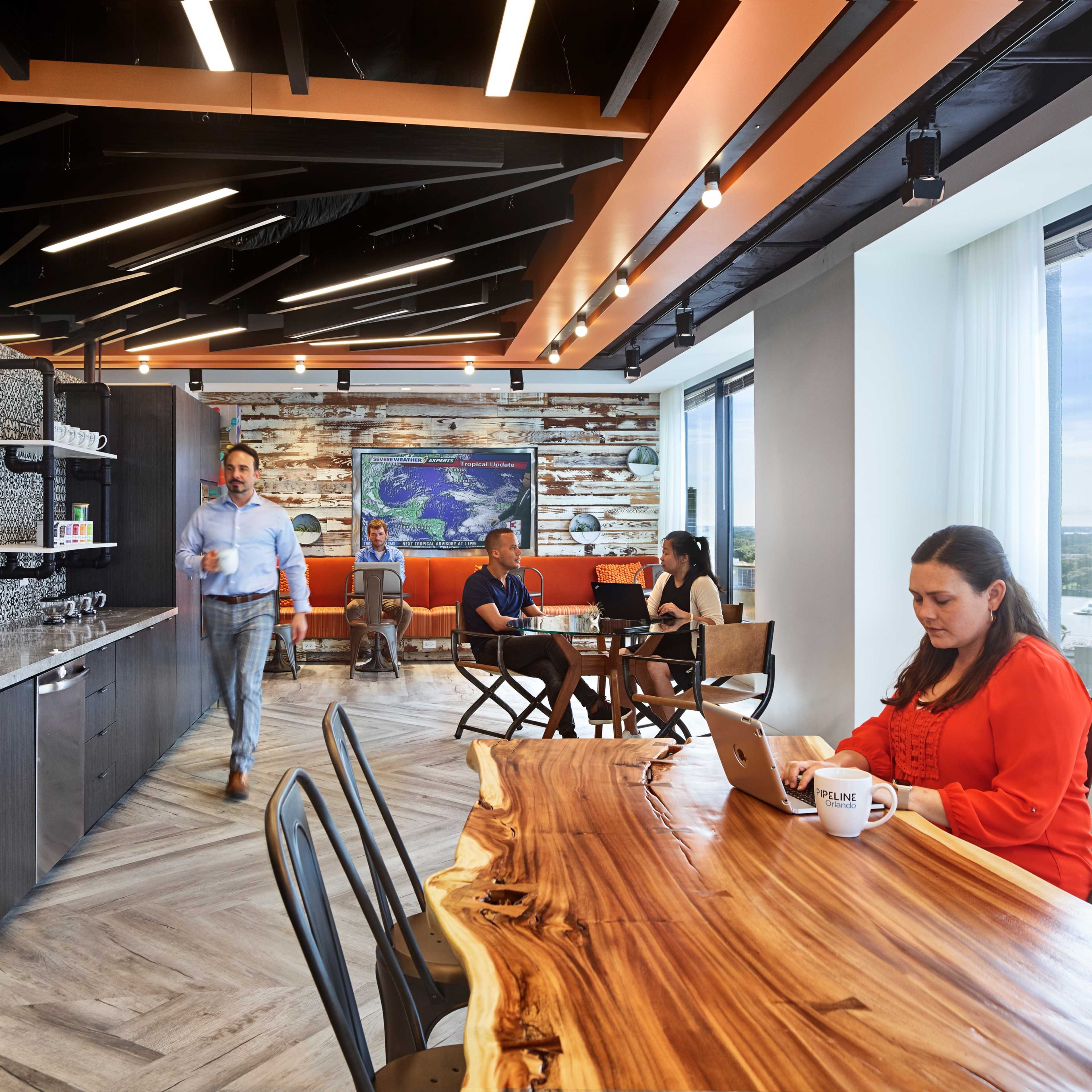 A modern office break room features a large wooden table, a kitchenette, and various people engaged in conversation and working on laptops.
