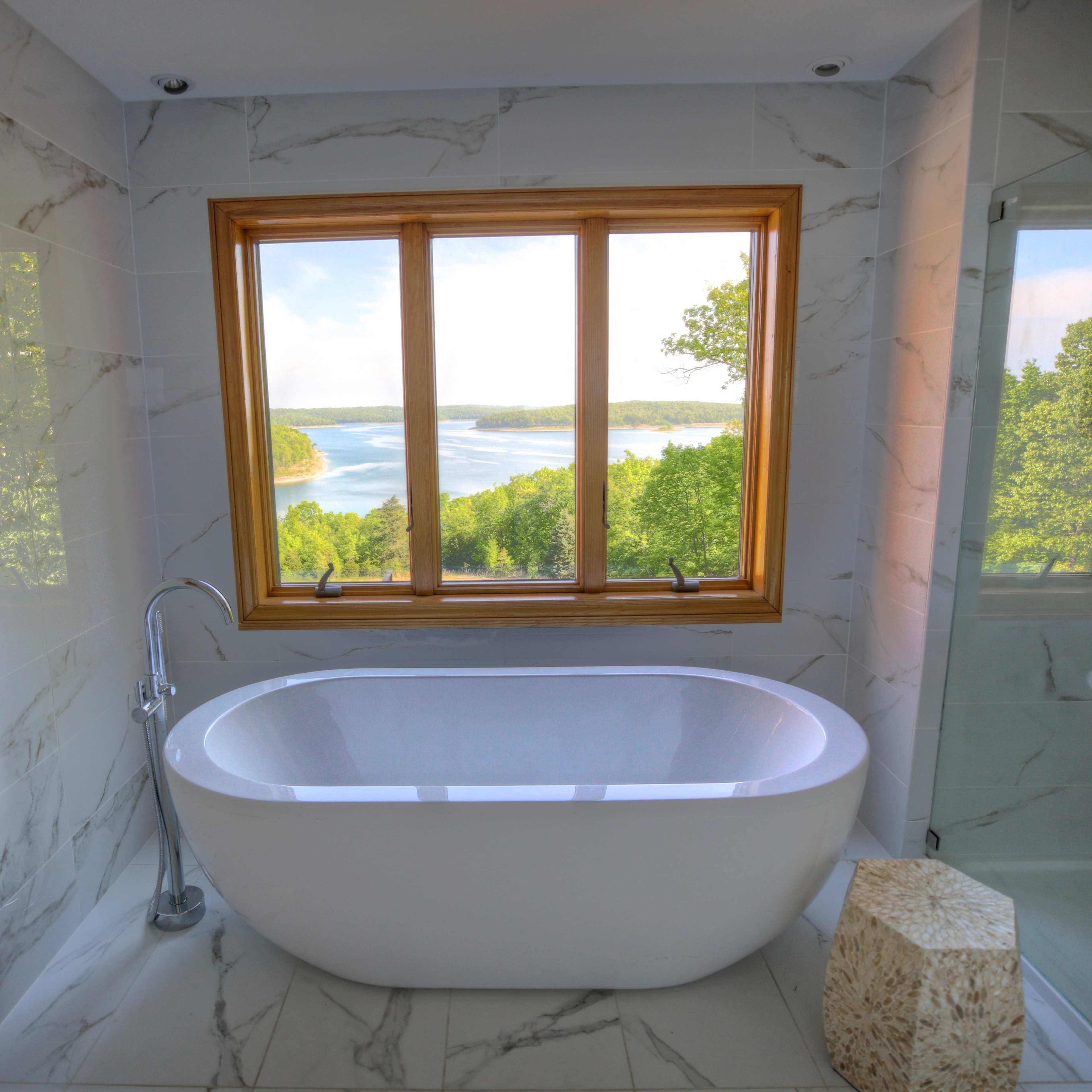 A modern bathroom features a freestanding bathtub near a large window with a view of a river and lush greenery.