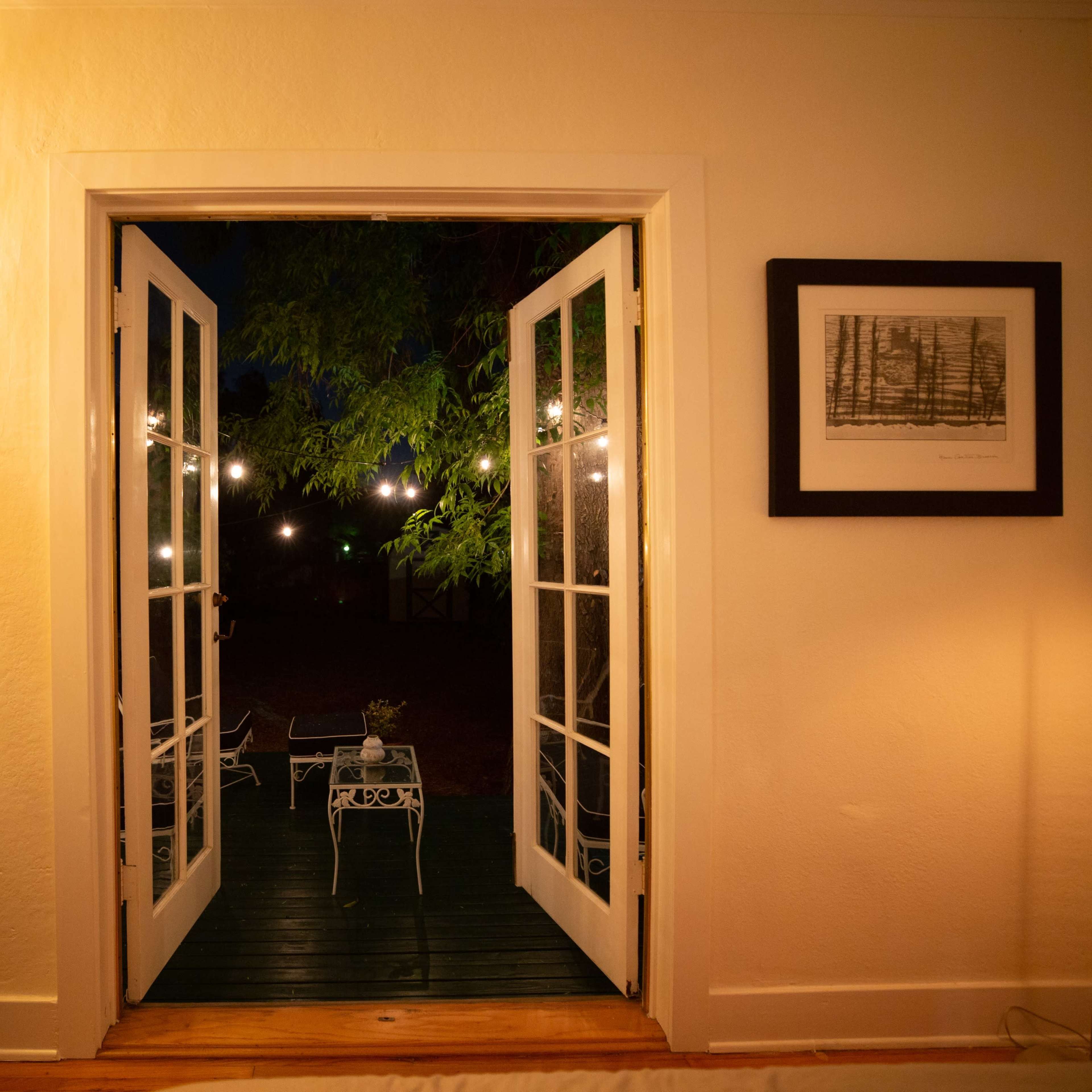 Open French doors leading to a porch illuminated by string lights, with a view of a garden setting.