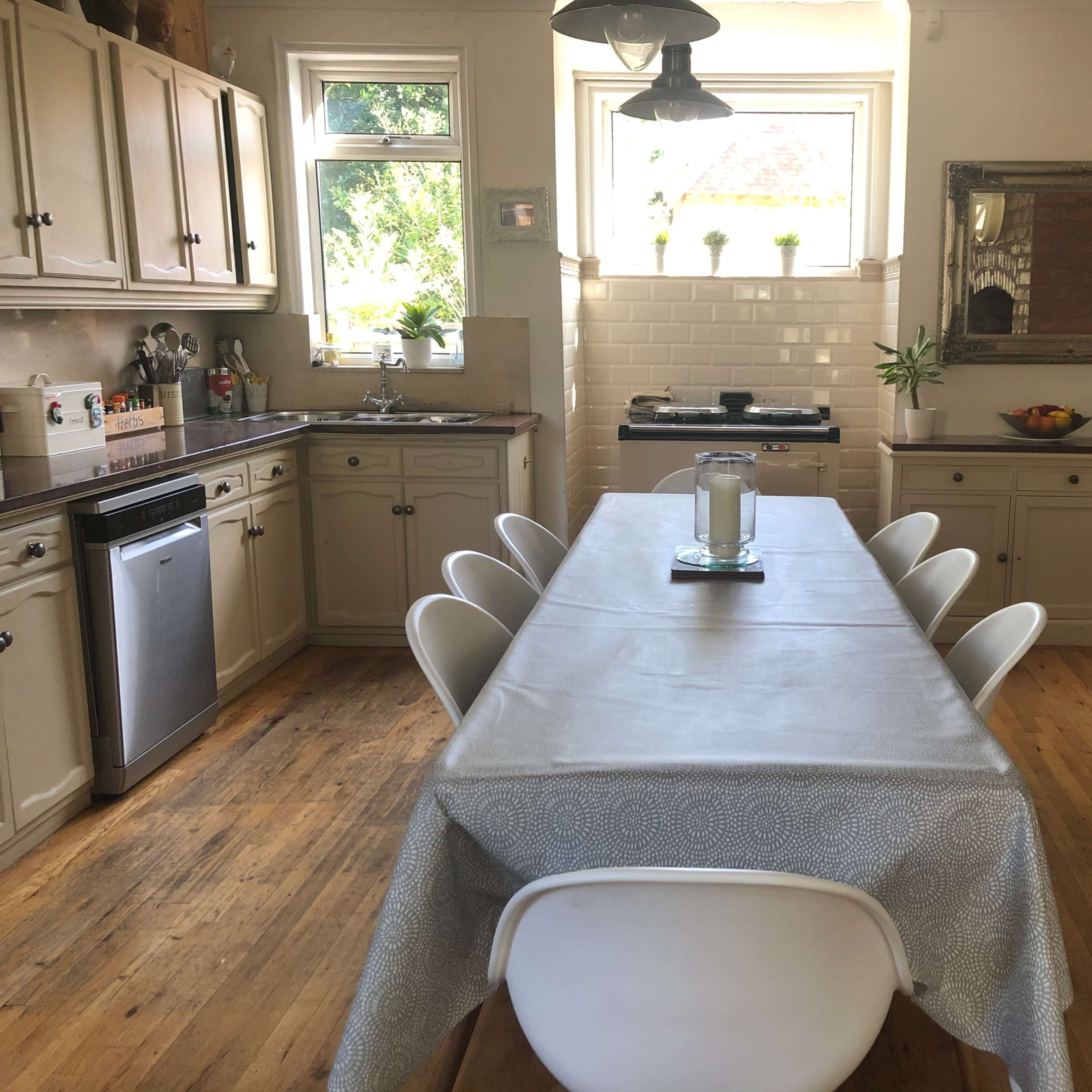 A spacious kitchen features a long dining table with white chairs, wooden floors, and cabinets along the walls, illuminated by natural light from the windows.