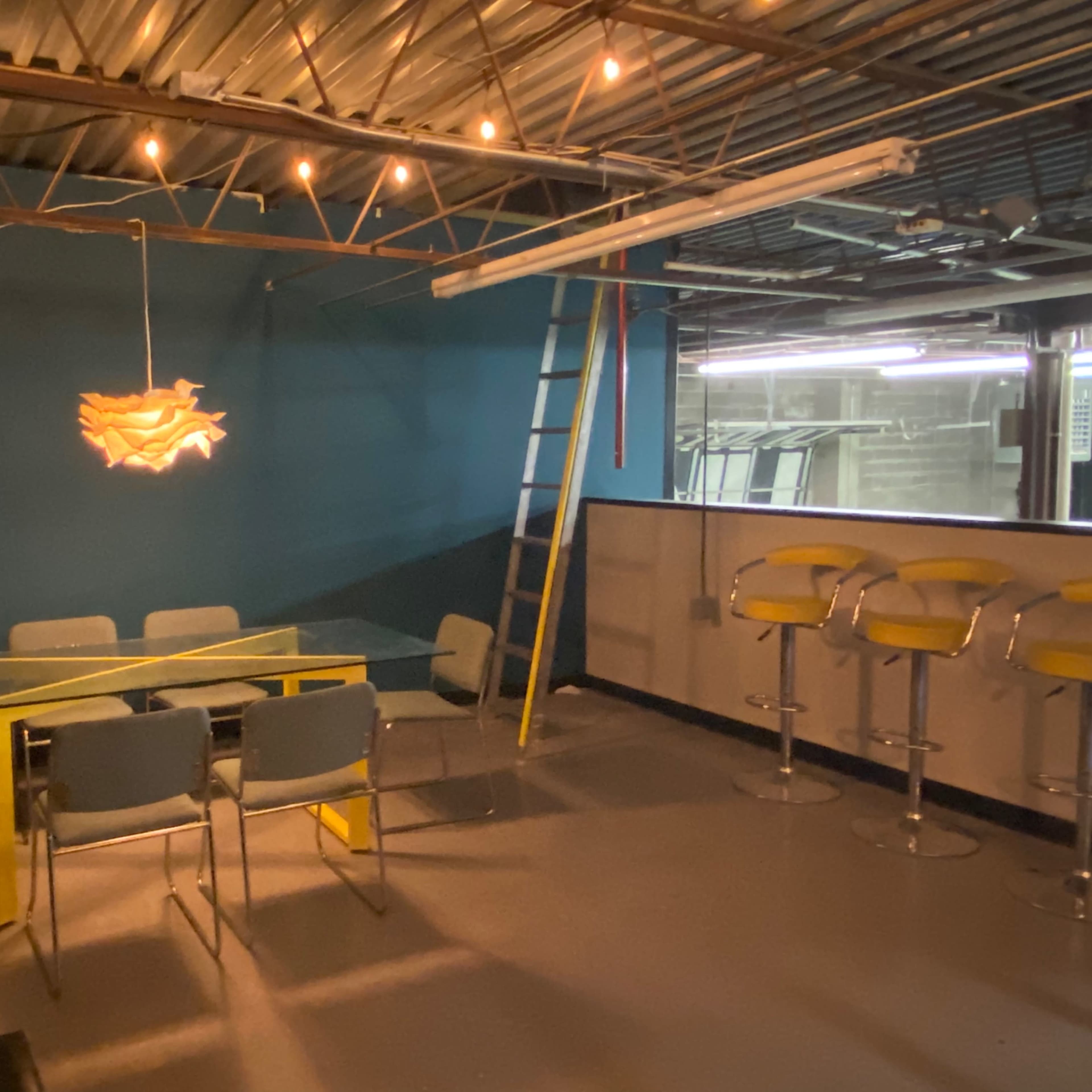 A modern meeting room with a glass table surrounded by six chairs, a yellow hanging light fixture, a bar area with tall stools, and a ladder leaning against the wall.