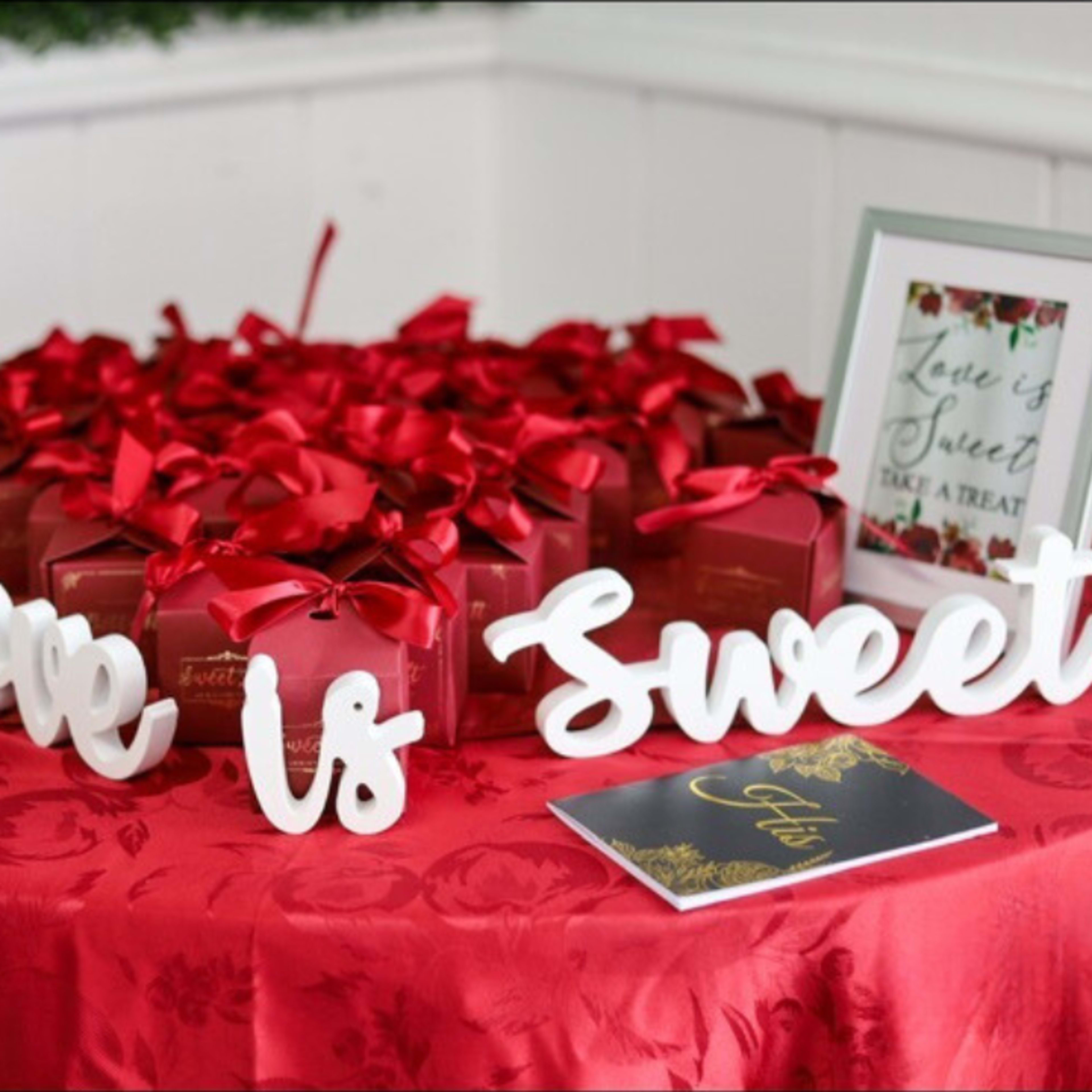 A table is decorated with a red tablecloth, featuring gift boxes tied with ribbons, framed signs, and a decorative message that reads "Love Is Sweet."