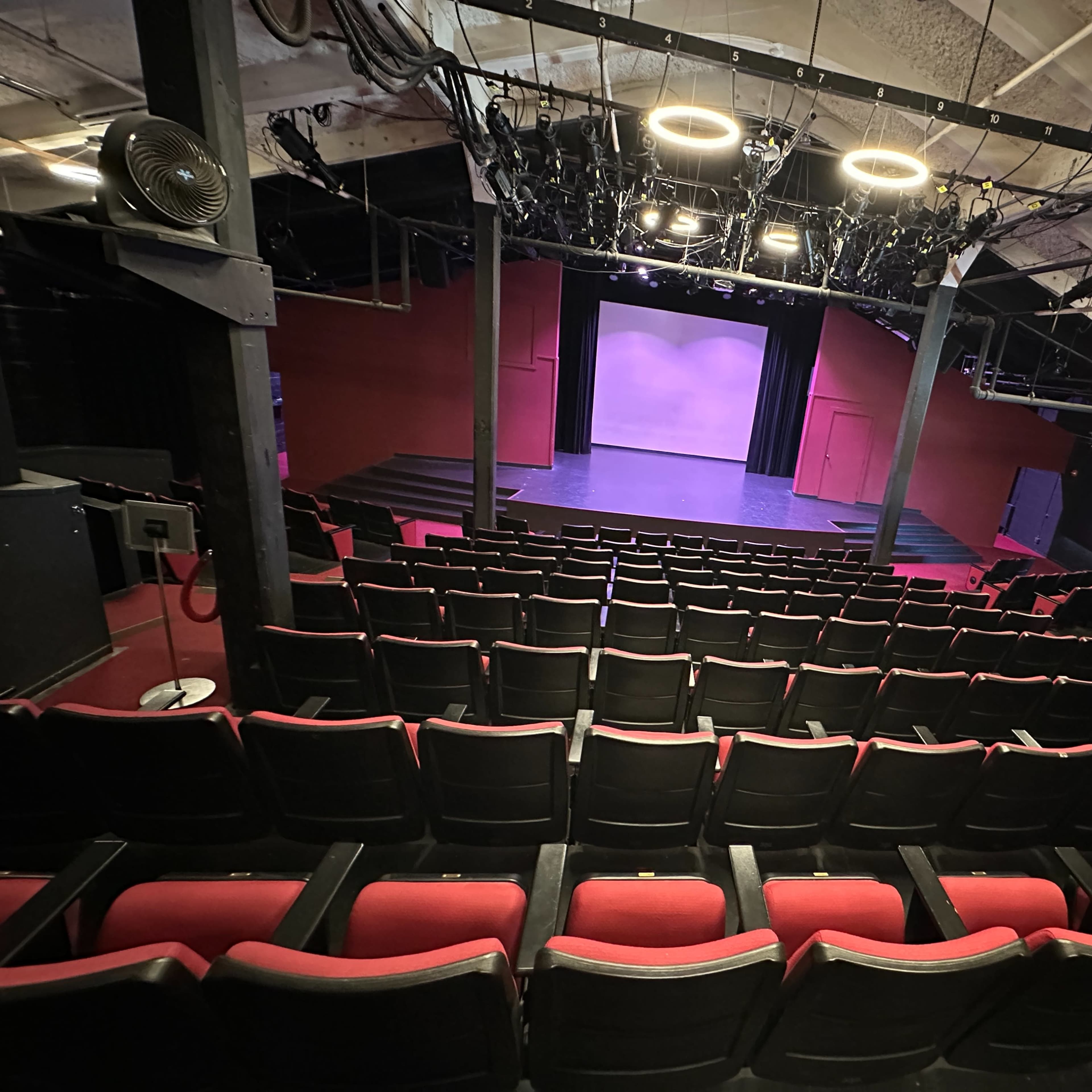 A theater with red seats facing a stage that is illuminated by overhead lights and has a purple backdrop.