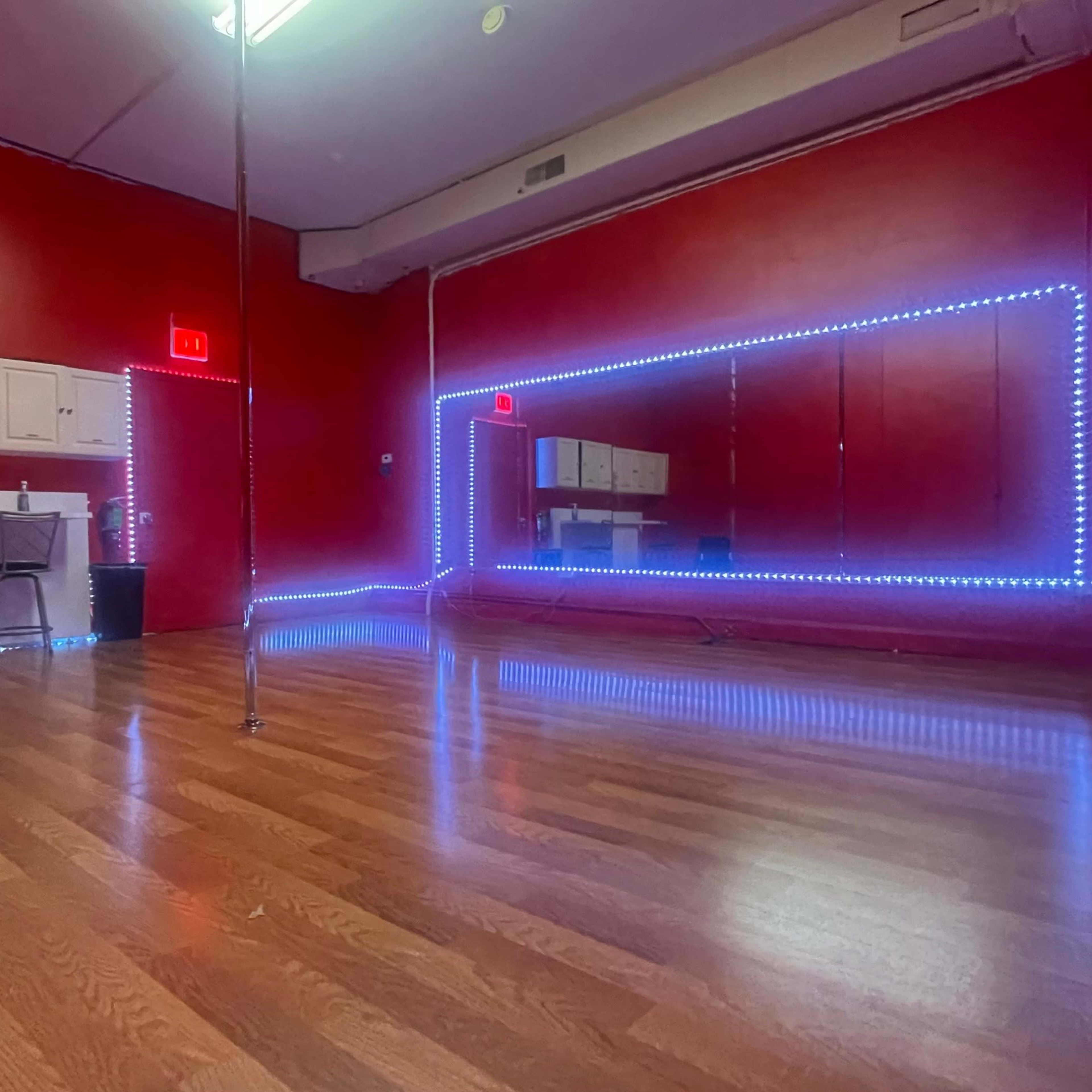 A dance studio features a polished wooden floor with red walls, illuminated by blue LED lights along the mirrors and perimeter.