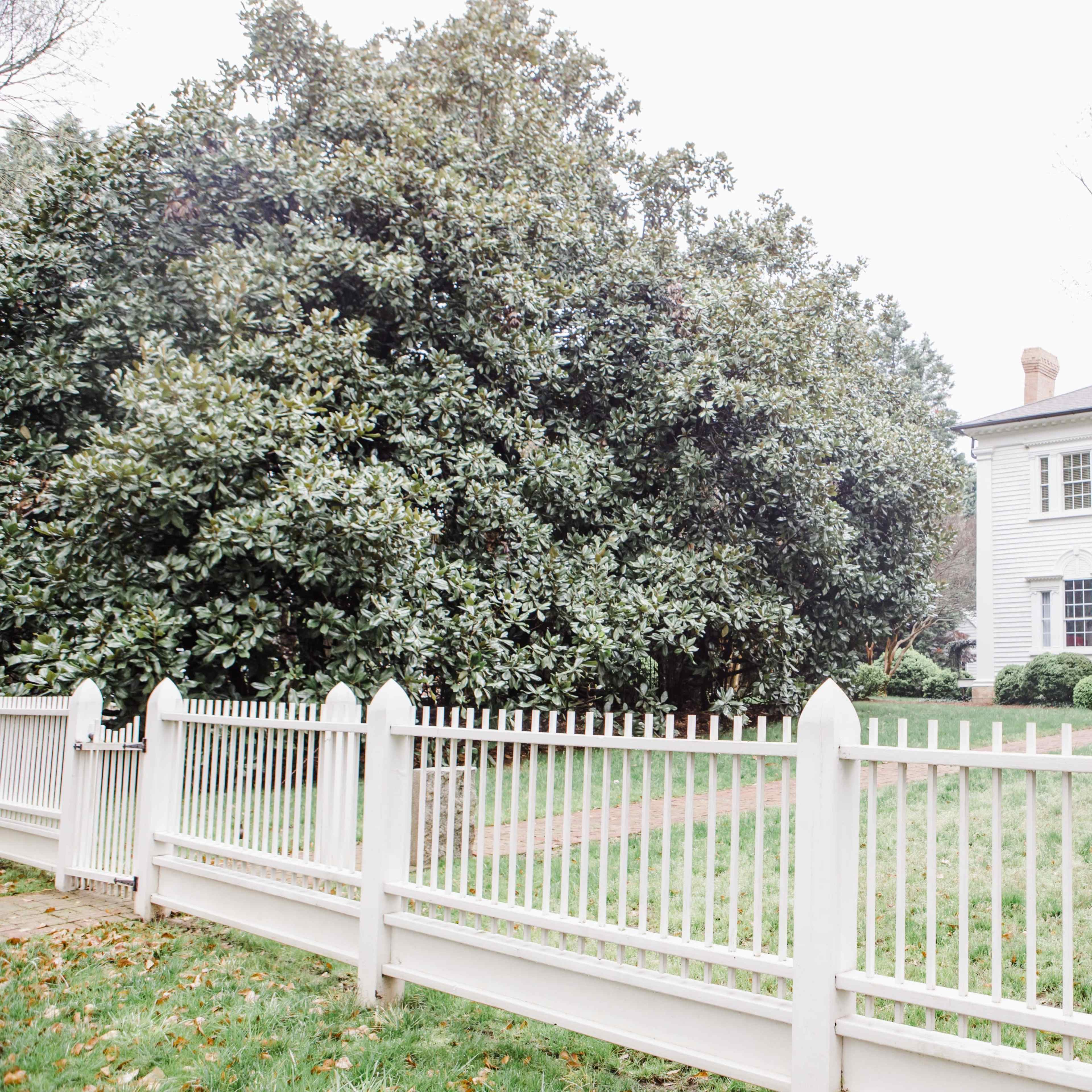 A large white house with a porch stands behind a white picket fence, surrounded by greenery.