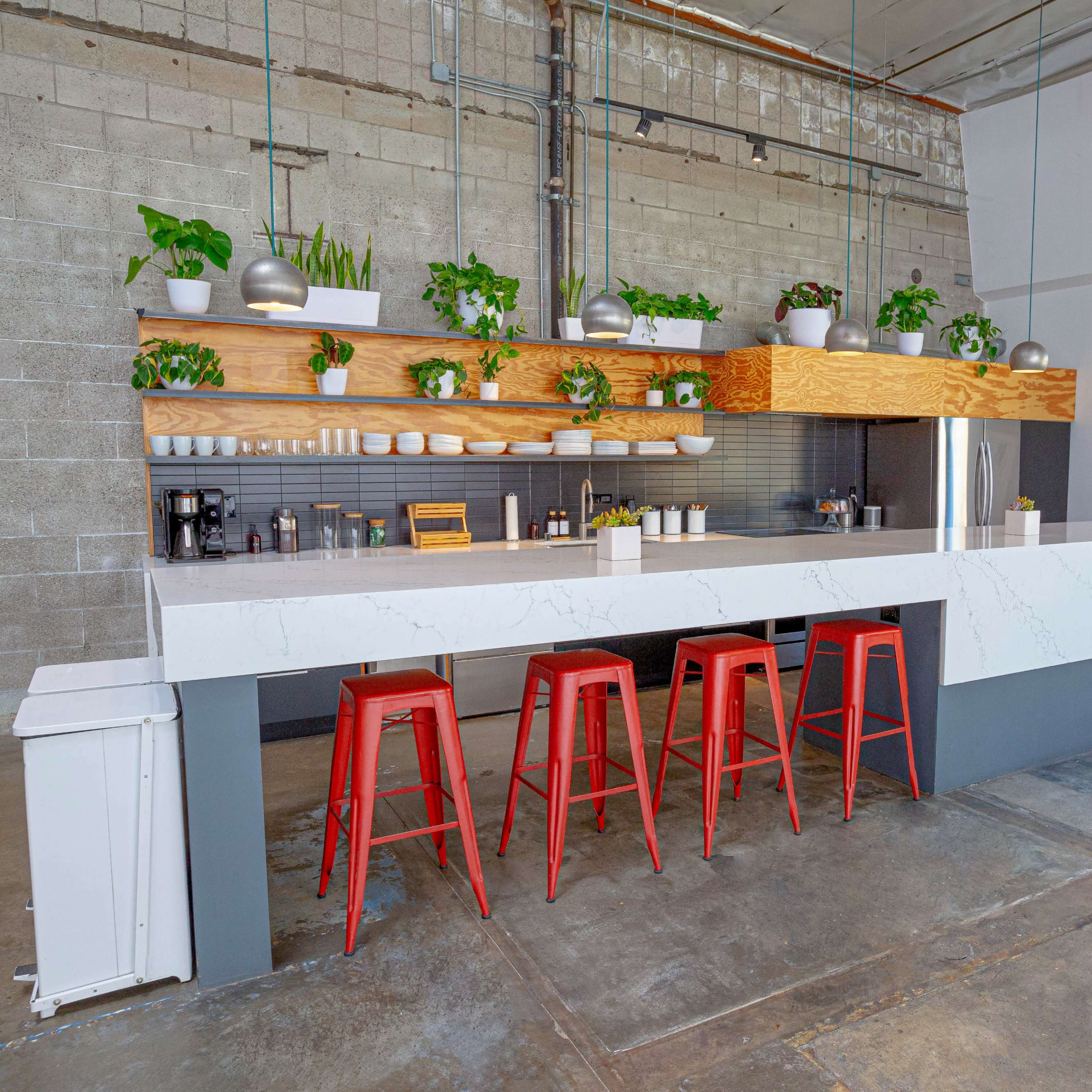 A modern kitchen bar with a white marble countertop, wooden shelving adorned with plants, and red metal stools.