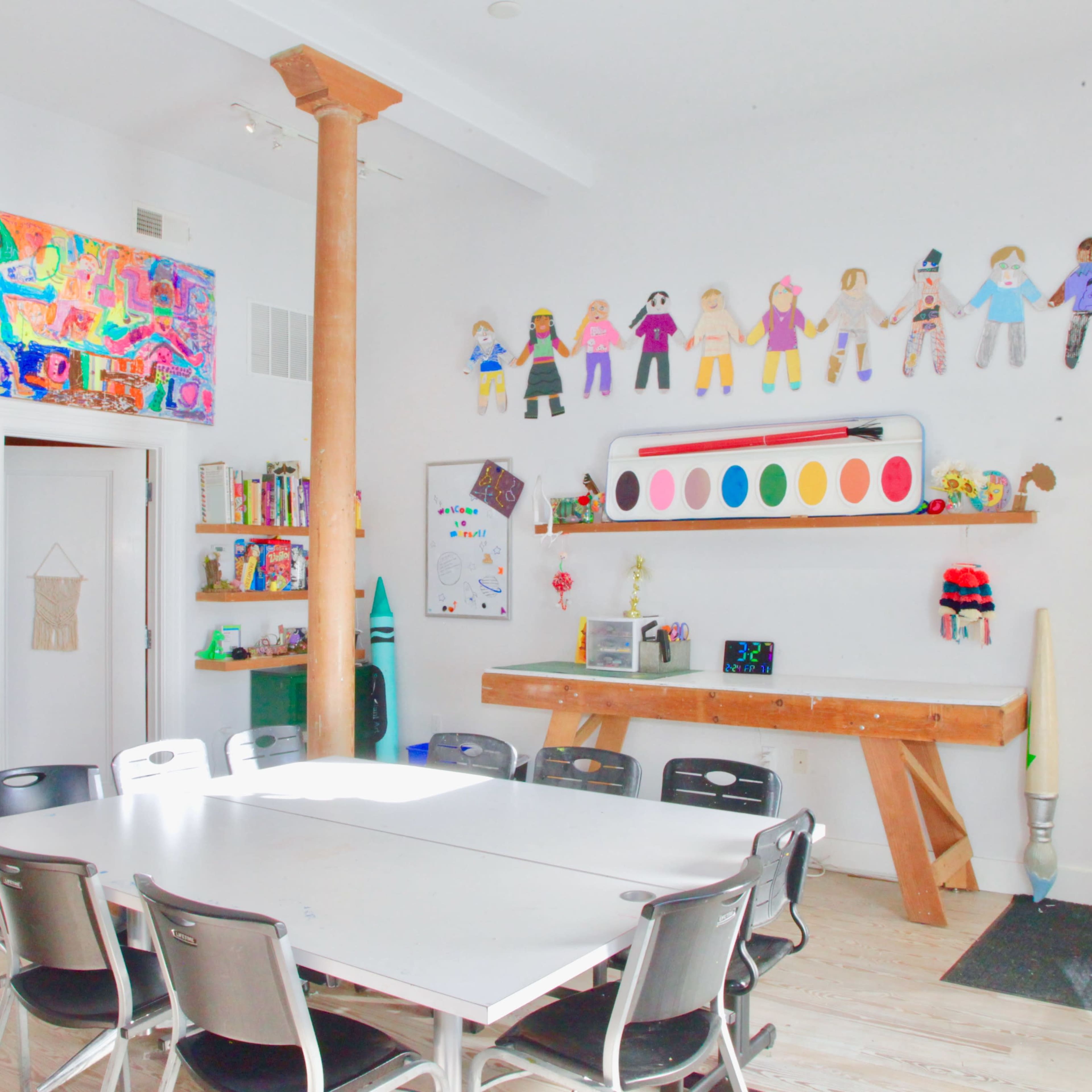 A brightly lit classroom with a large table surrounded by chairs, colorful student artworks on the walls, and various teaching materials on shelves.