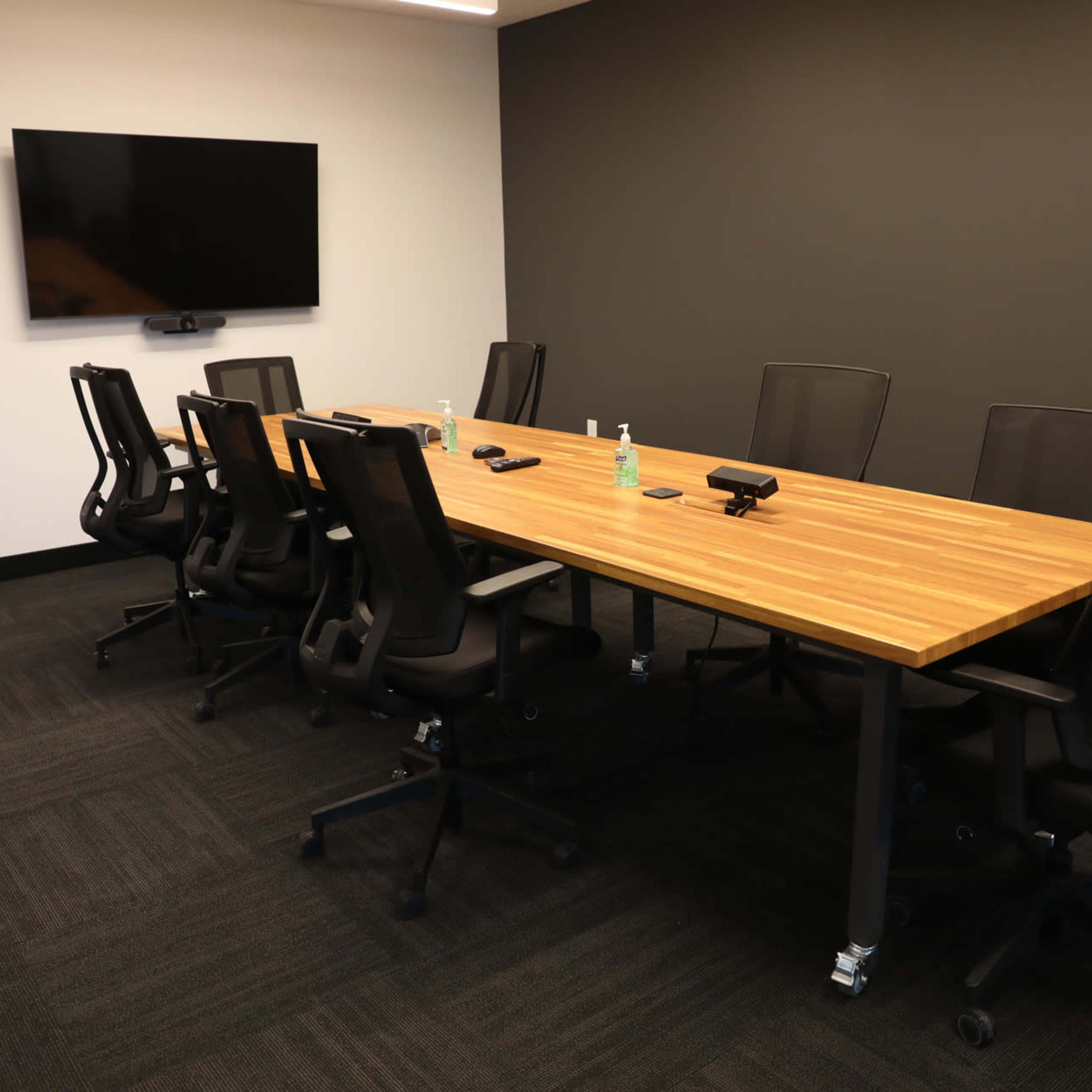 A conference room features a large wooden table surrounded by eight black office chairs, with a television mounted on the wall.