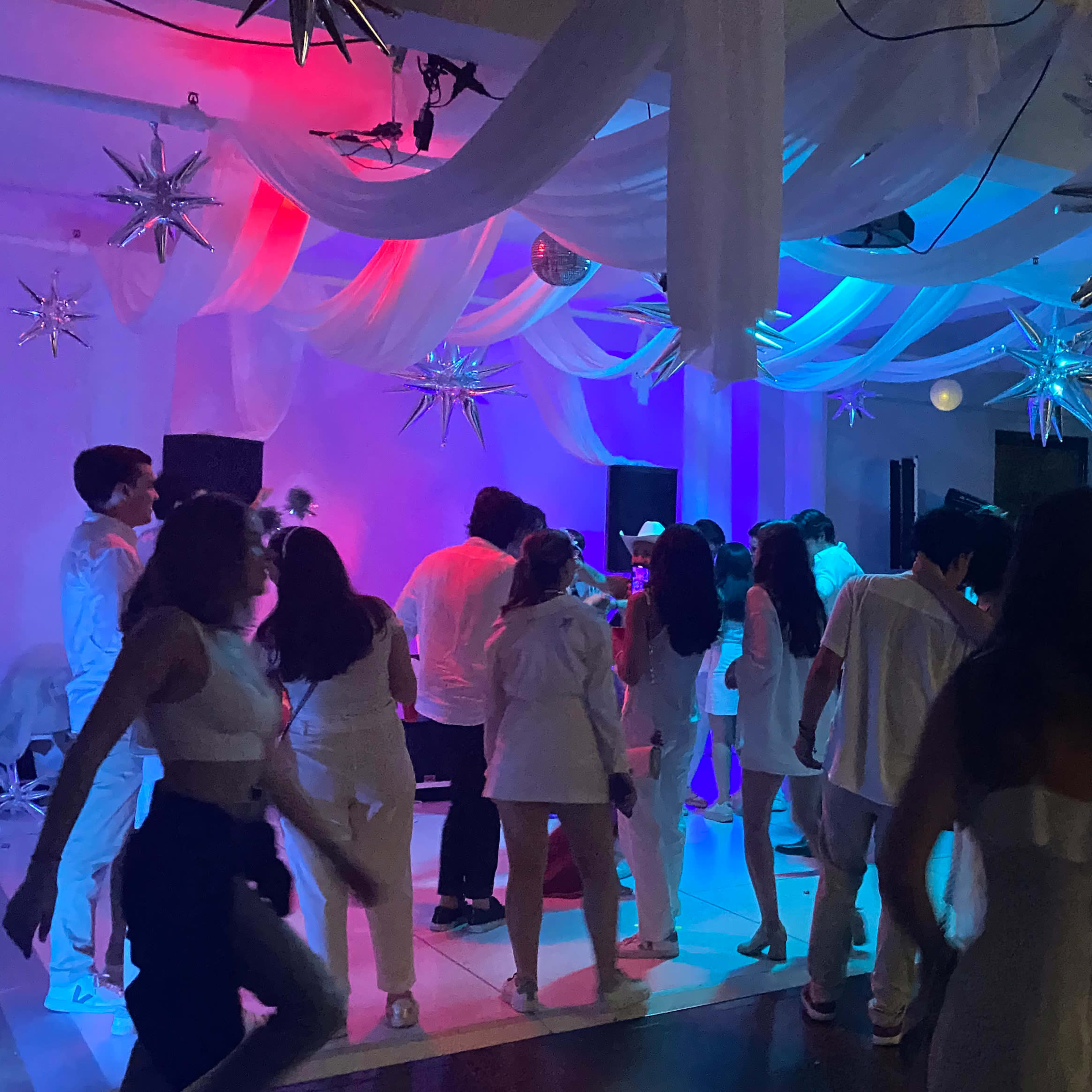 A group of people dressed in white are dancing at an event illuminated by colorful lights with decorative stars hanging from the ceiling.