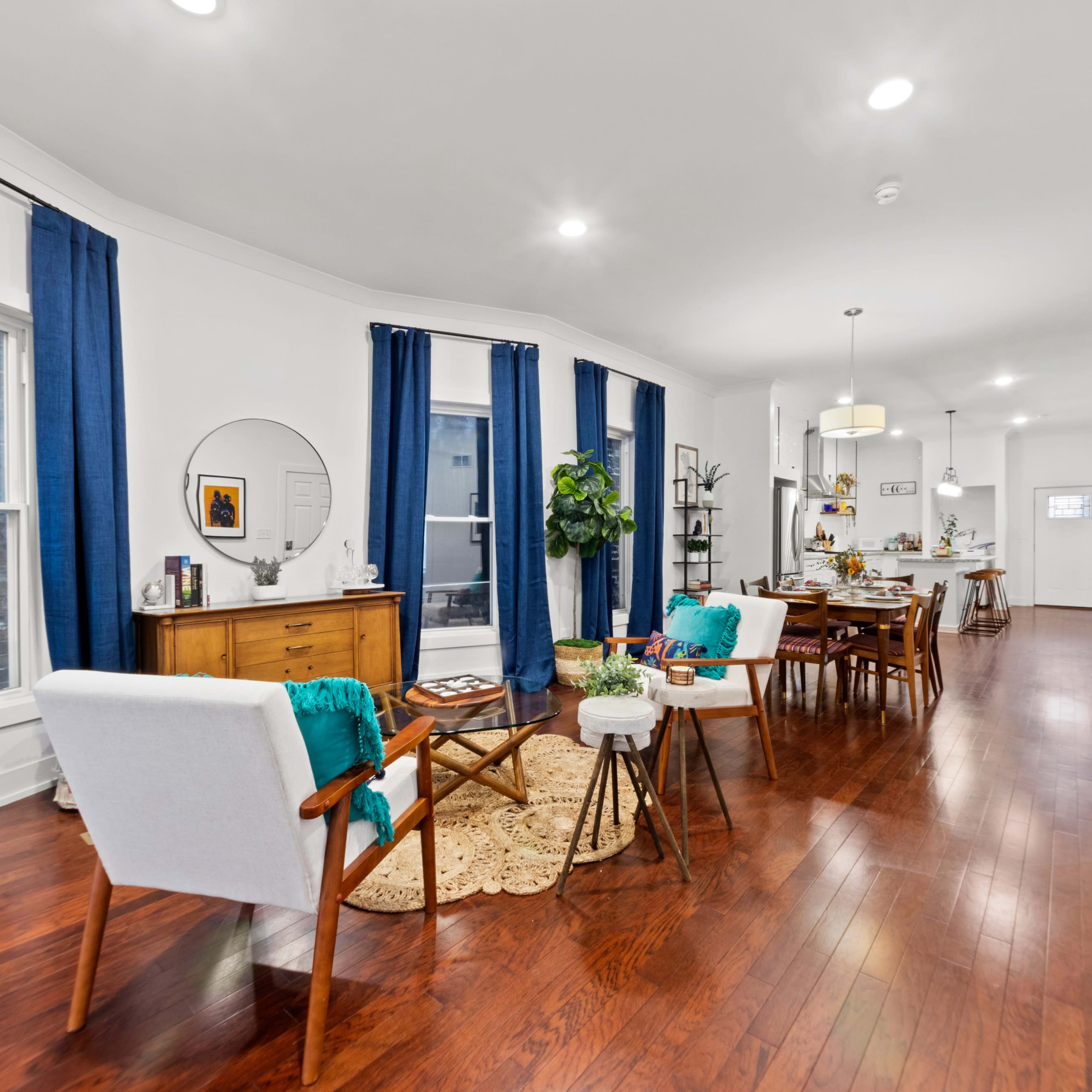 A modern living space features hardwood floors, white walls, and a combination of a dining area and lounge with blue curtains, wooden furniture, and decorative plants.