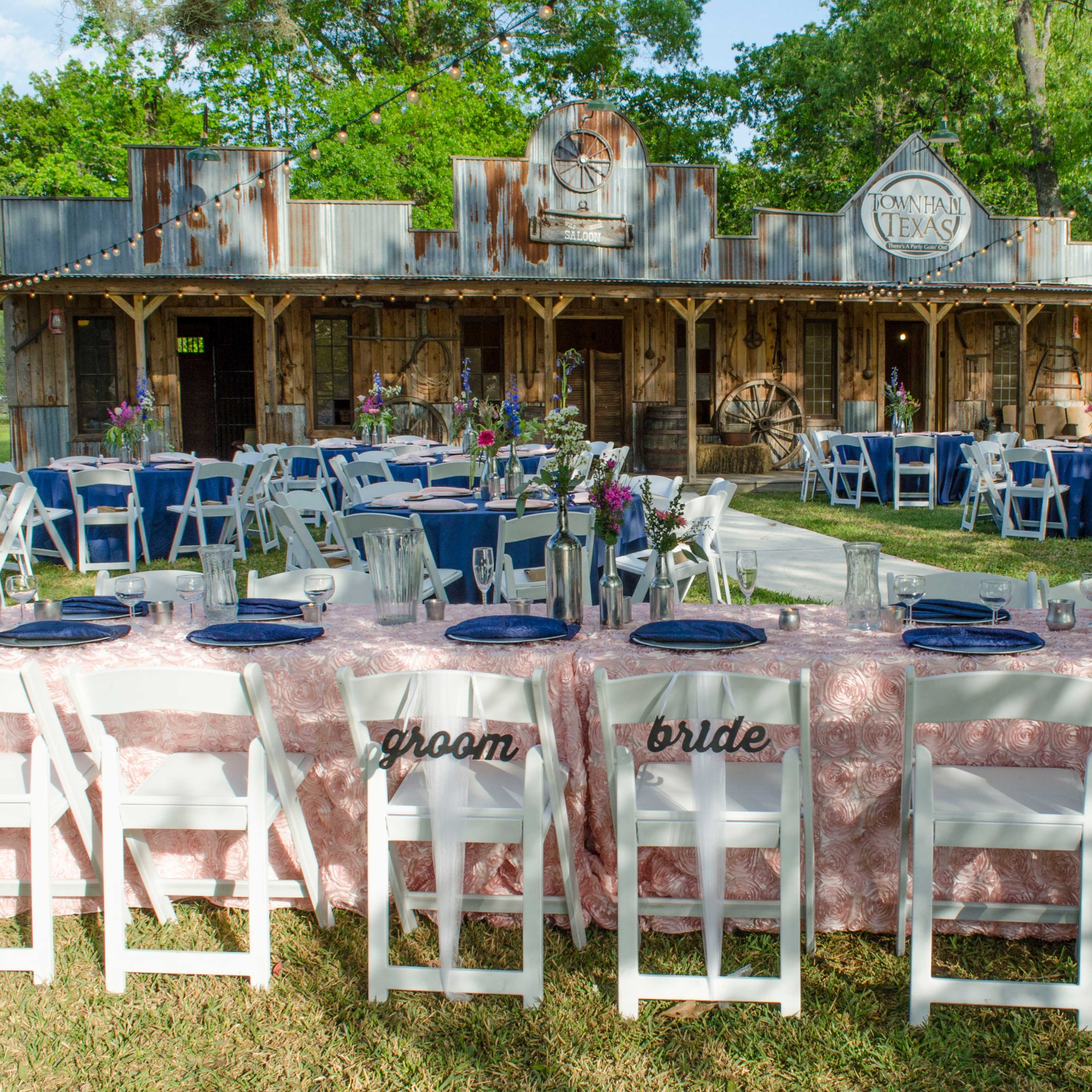 A outdoor event setup features tables with white chairs and blue table settings in front of a rustic building with a weathered facade.