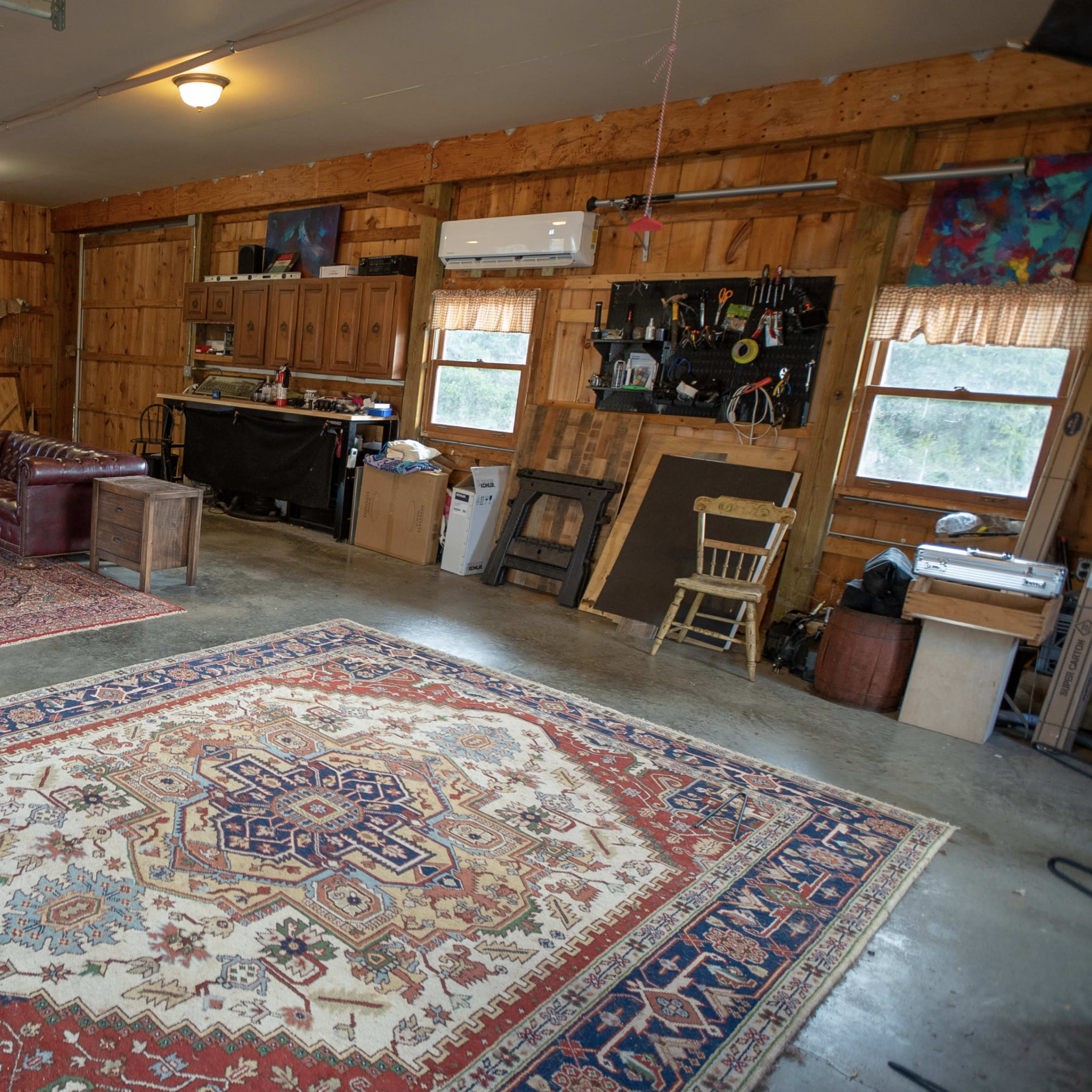 A spacious garage with wooden walls, featuring an American flag, a carpeted area with a large patterned rug, a couch, and various tools and furniture along the sides.
