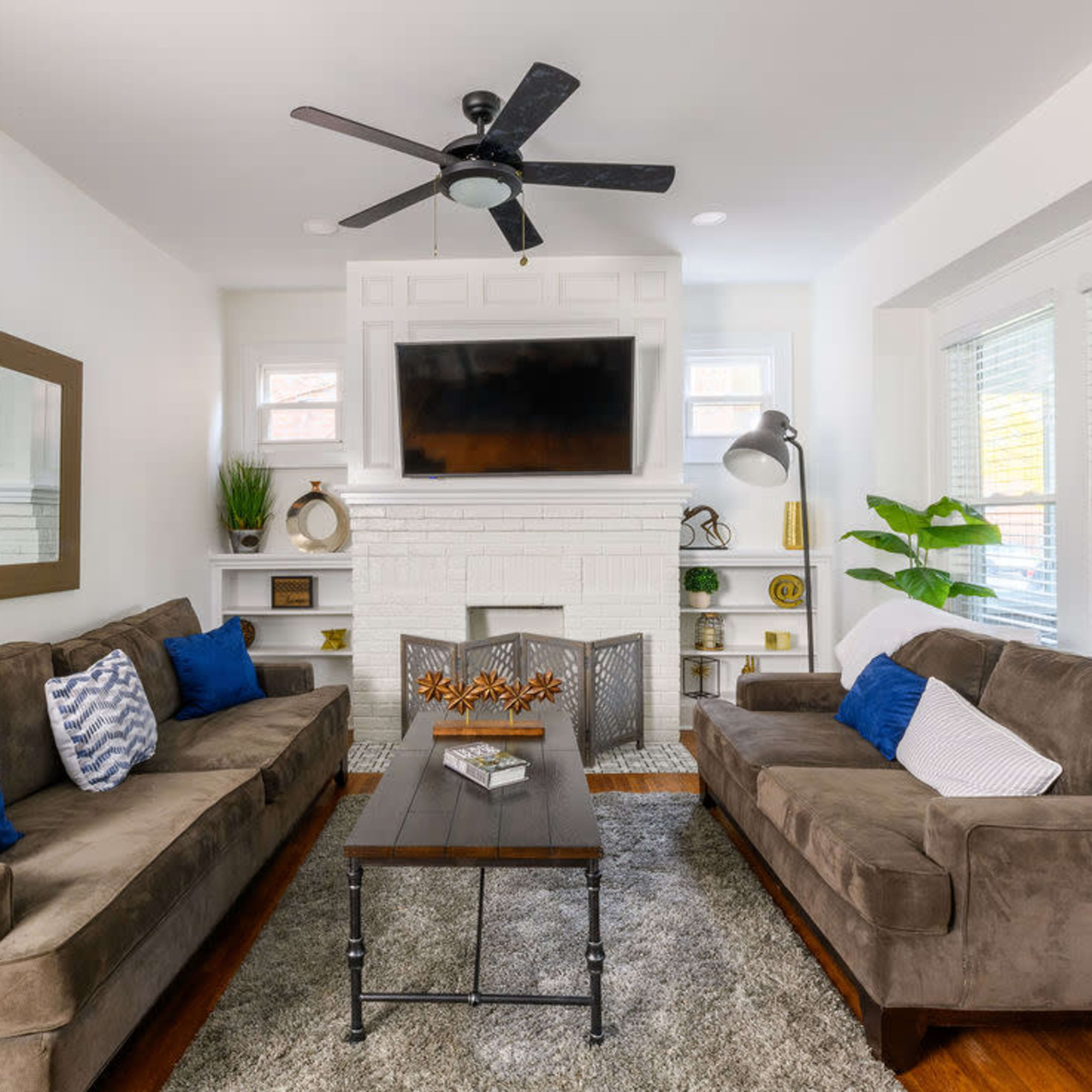 A living room features two brown sofas with blue and white pillows, a coffee table in the center, and a television mounted above a white brick fireplace, with large windows allowing natural light.