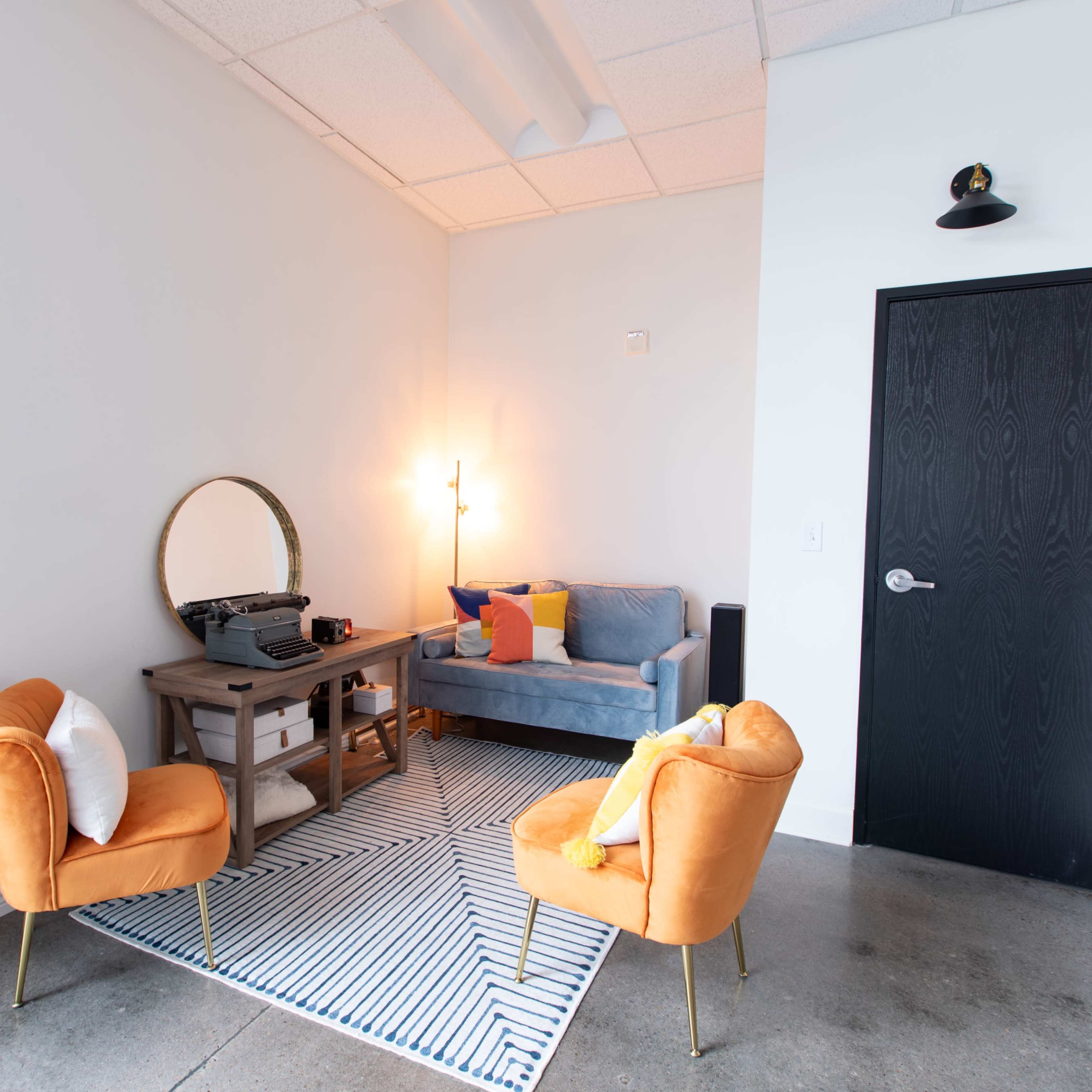 A small, modern lounge area with two orange chairs, a gray sofa, a round mirror, and a vintage typewriter on a table, set against white walls and a concrete floor.