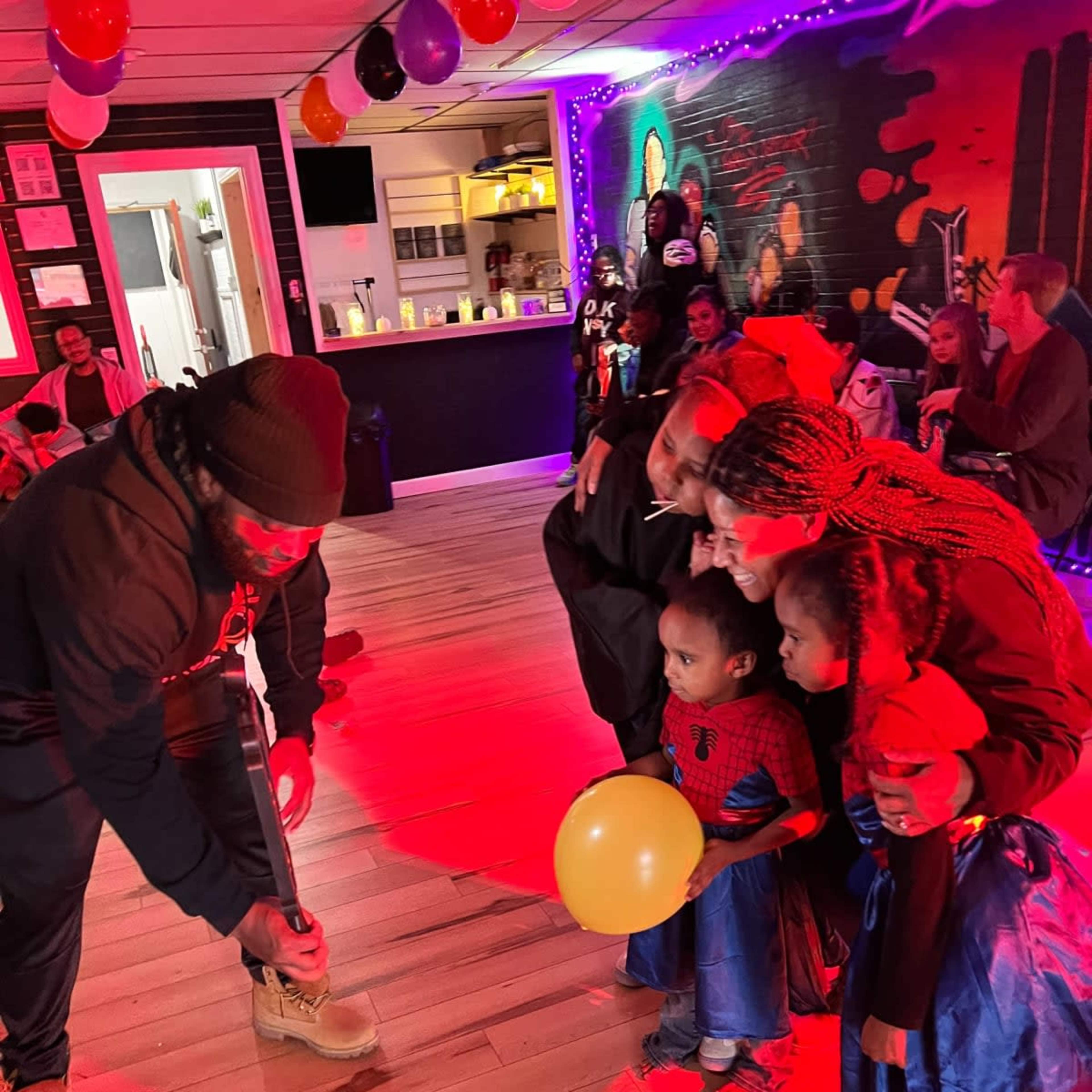 A man interacts with children in costumes during a festive event illuminated by red lights.