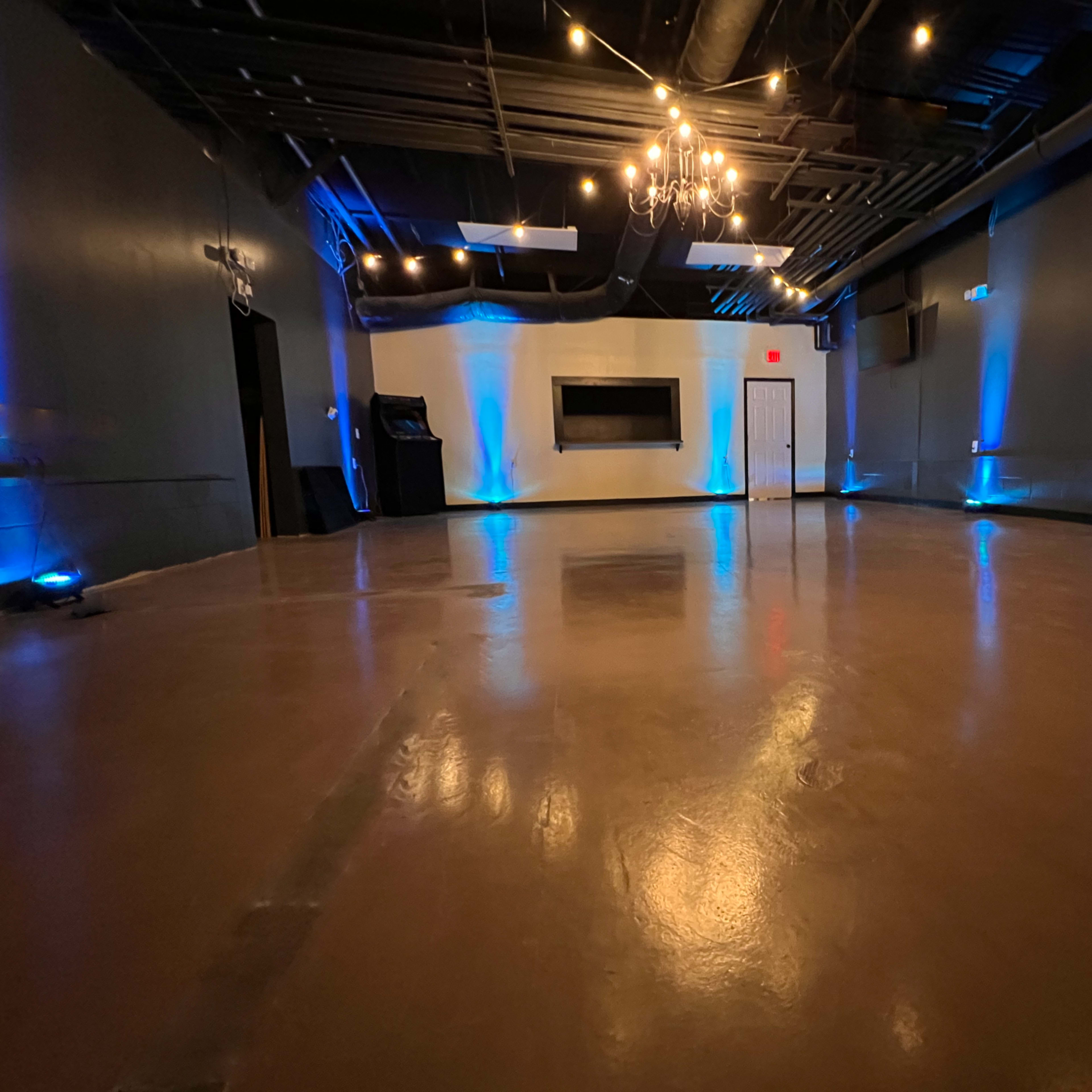 A spacious, empty event room with polished floors, blue lighting, and a chandelier.