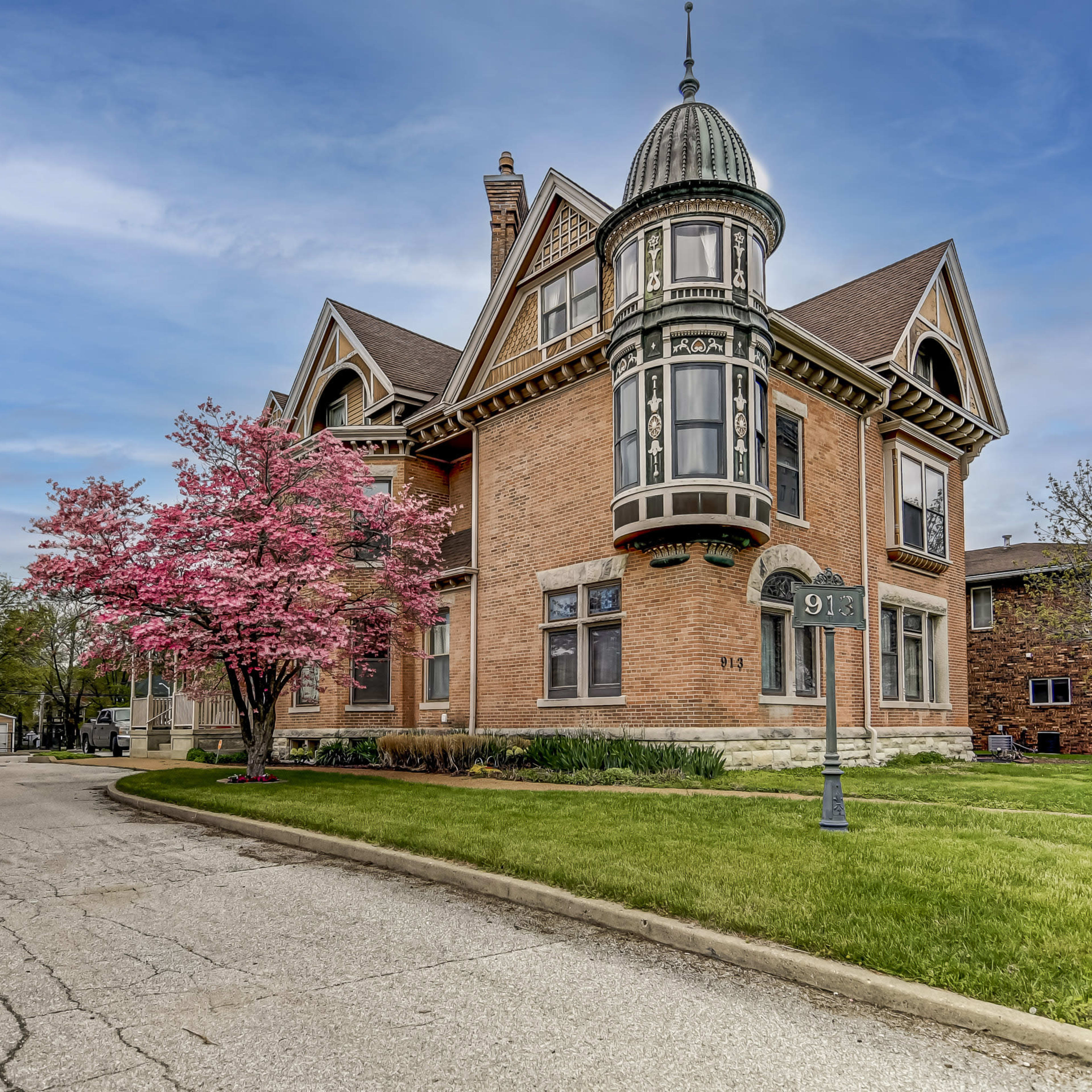 A large, historic brick house with a turret and a blooming pink tree in front, situated on a quiet street.