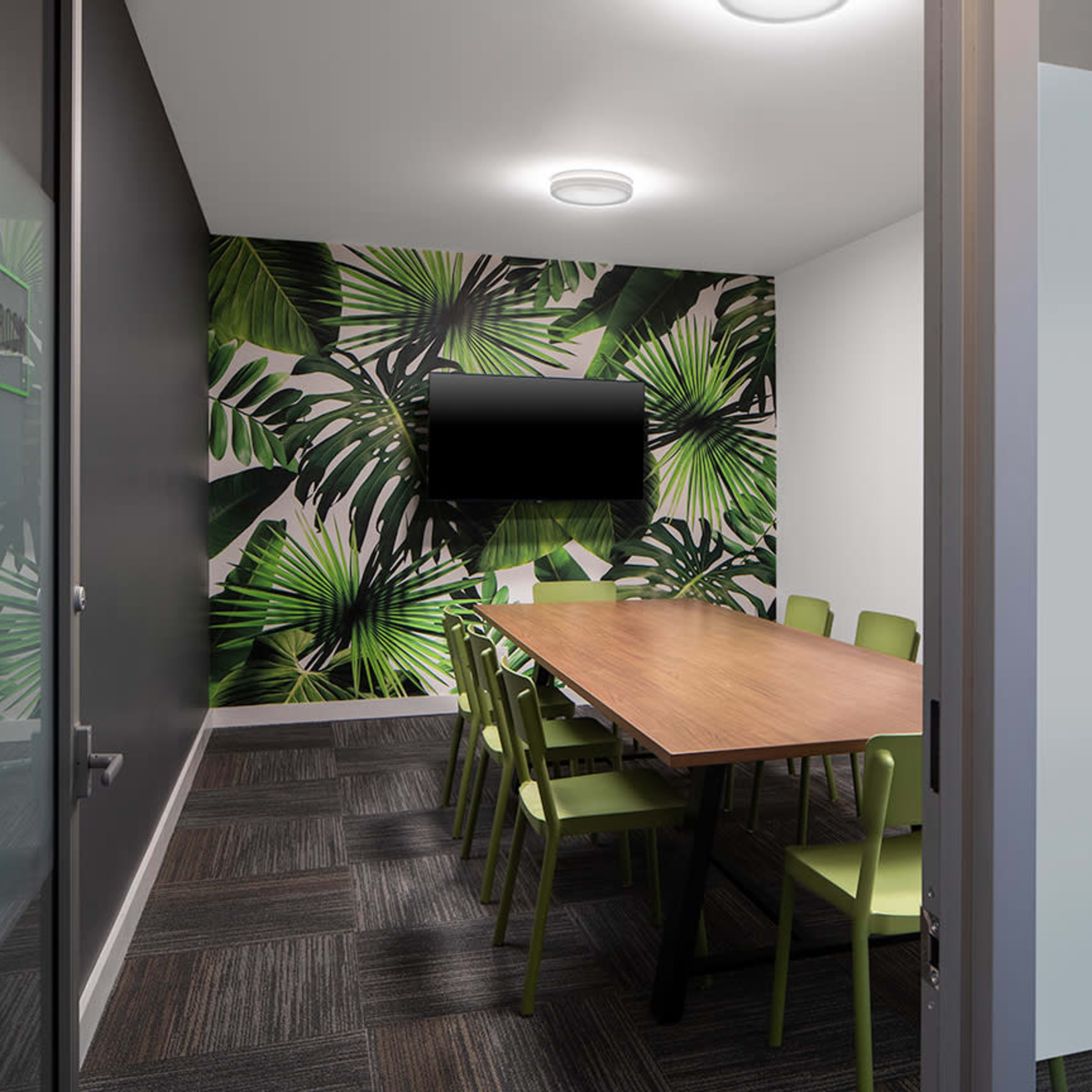 A conference room features a long wooden table surrounded by green chairs, with a wall decorated in tropical leaf patterns and a wall-mounted TV.