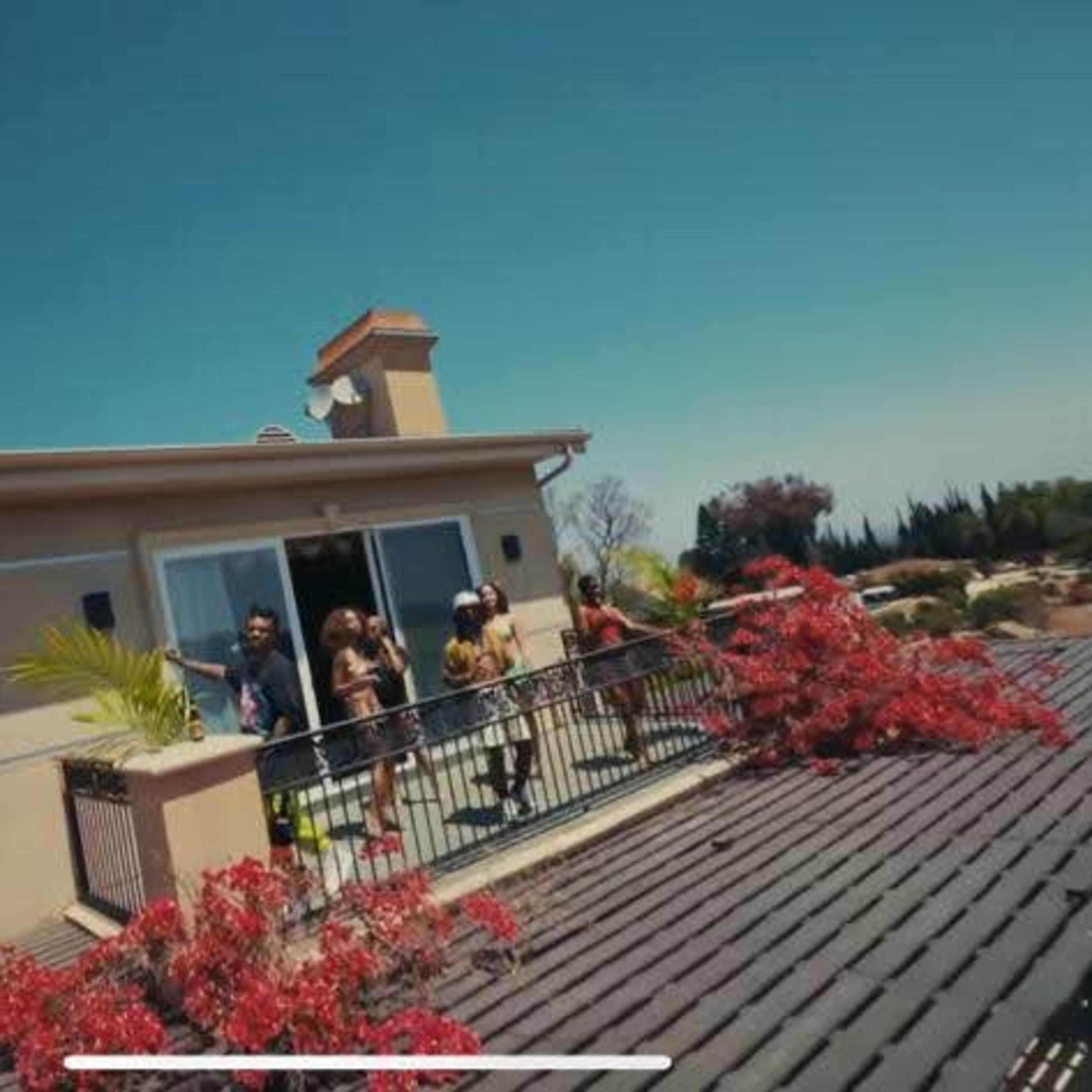 A group of people stands on a balcony of a house surrounded by greenery and vivid flowers under a clear blue sky.