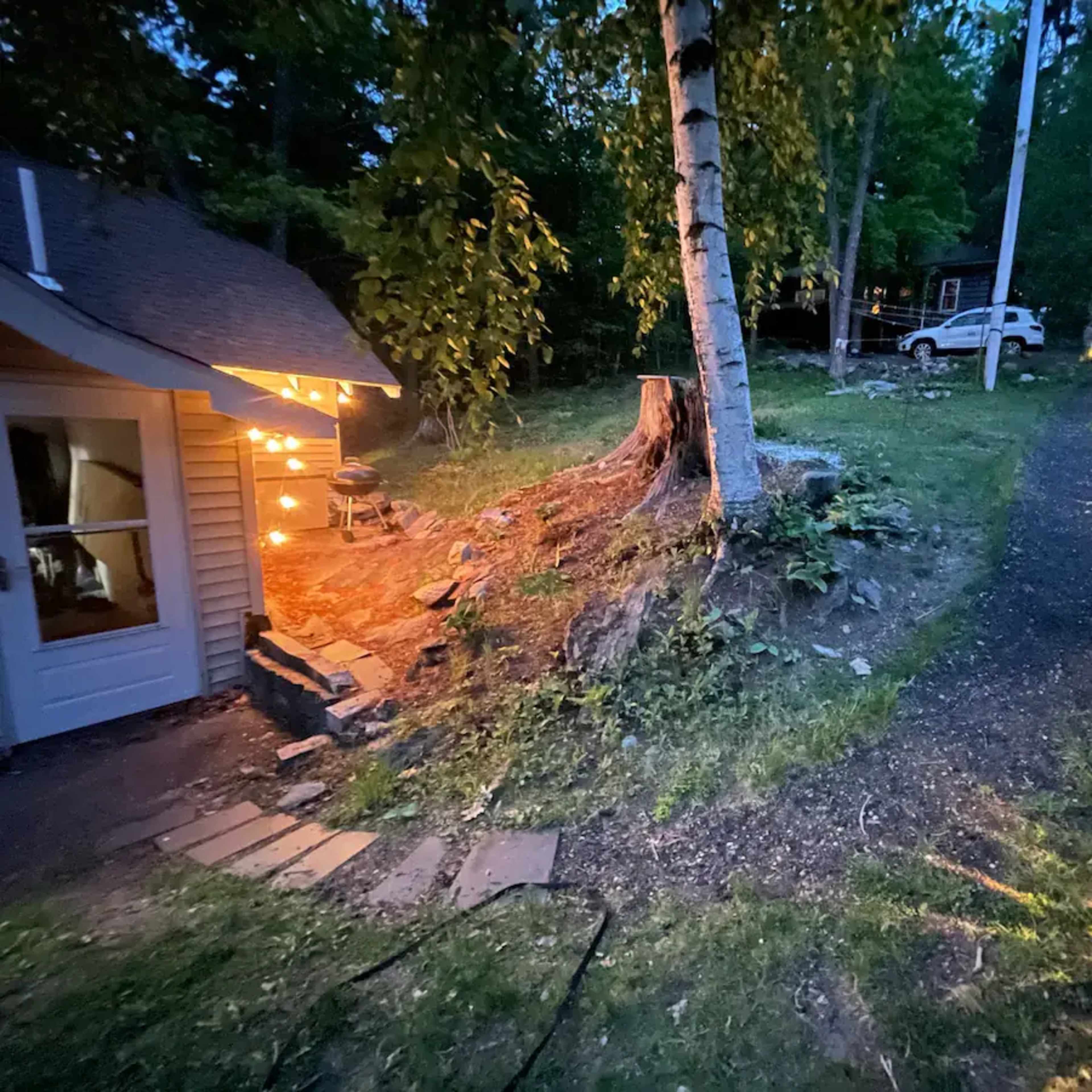 A path winds through a wooded area, with a tree stump and a cabin illuminated by string lights on one side and a house visible in the distance on the other.