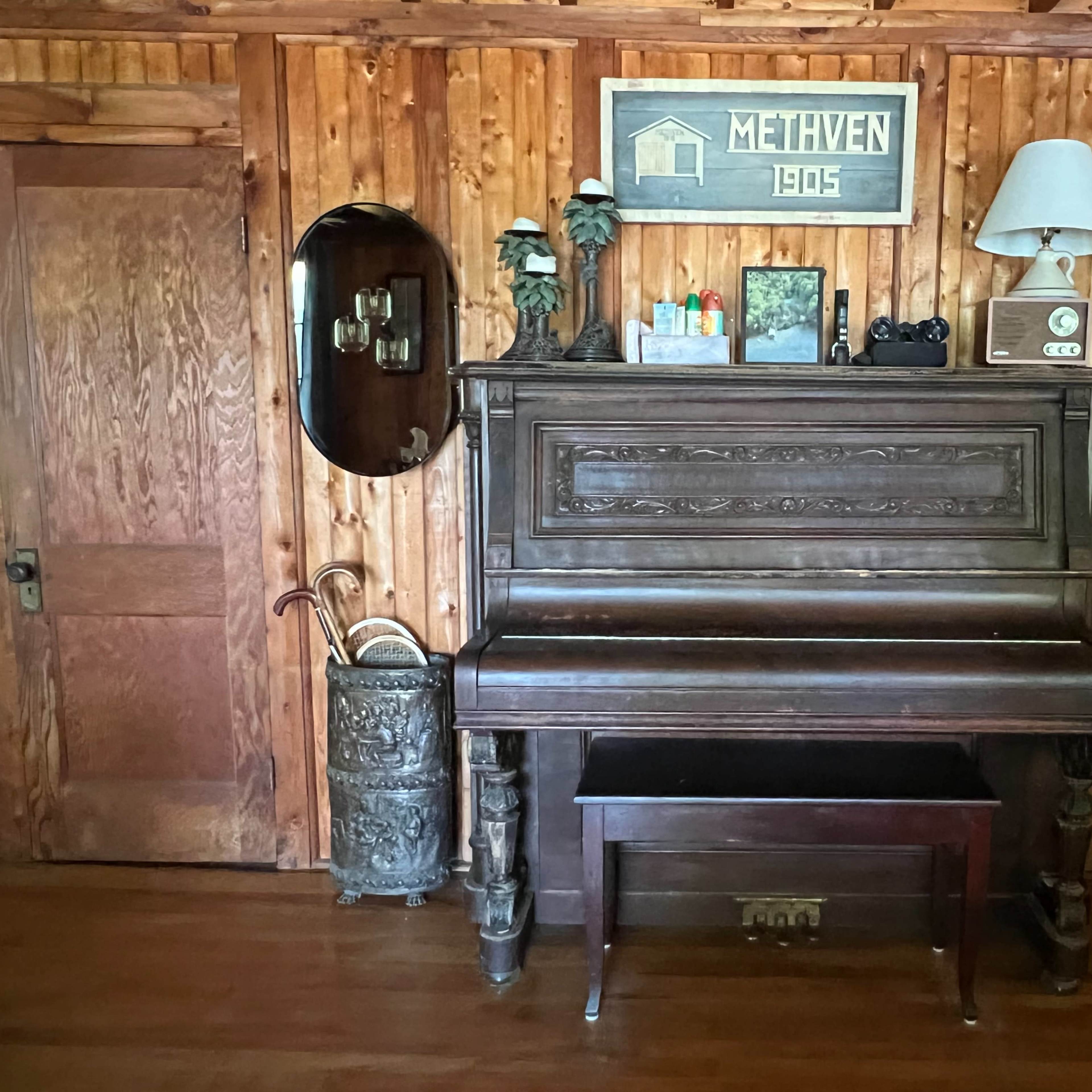 A vintage piano is positioned against a wooden wall, accompanied by a lamp, a mirror, and various decorative items on a shelf above it.