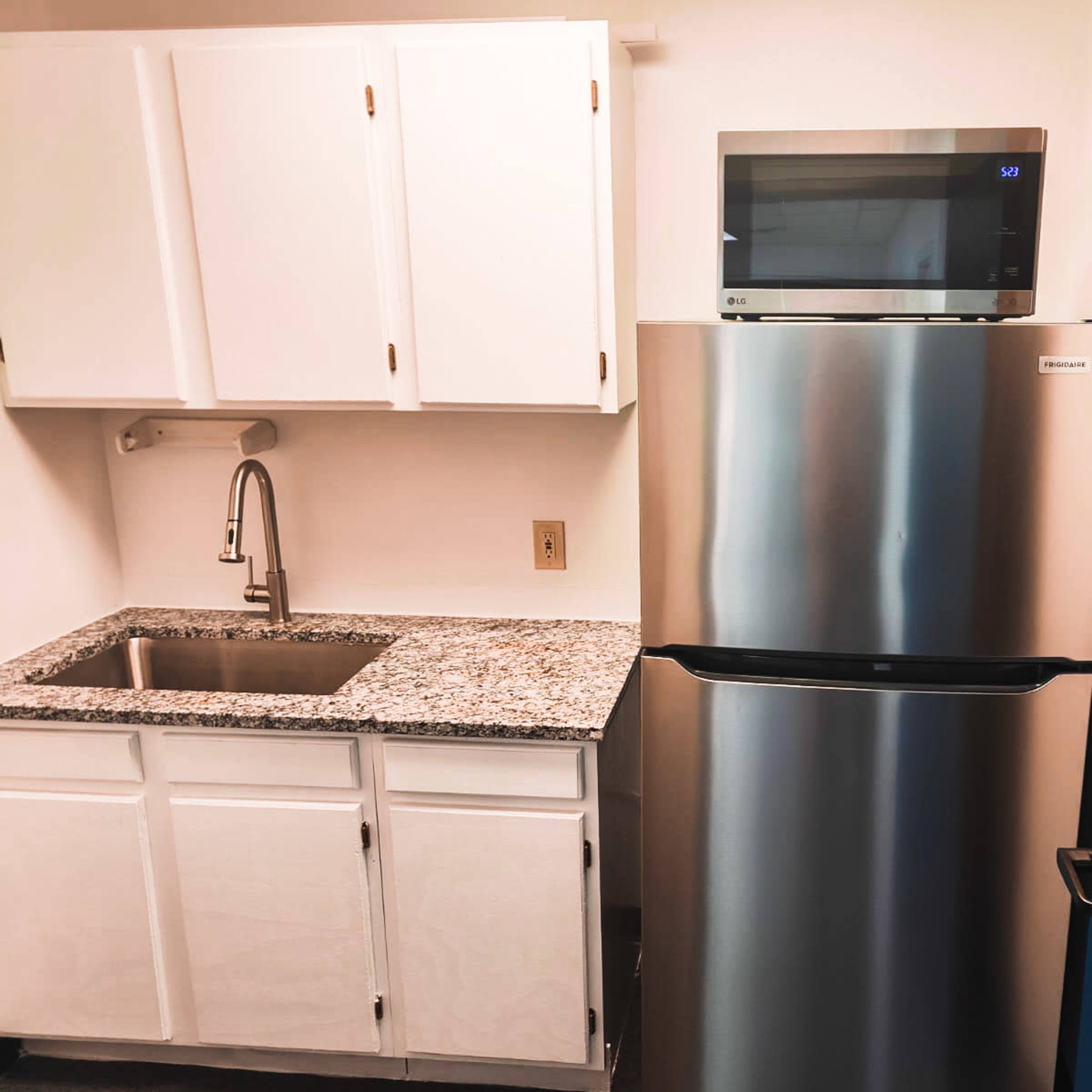 A kitchen area with a stainless steel refrigerator, a small microwave on top, a granite countertop, and white cabinets.