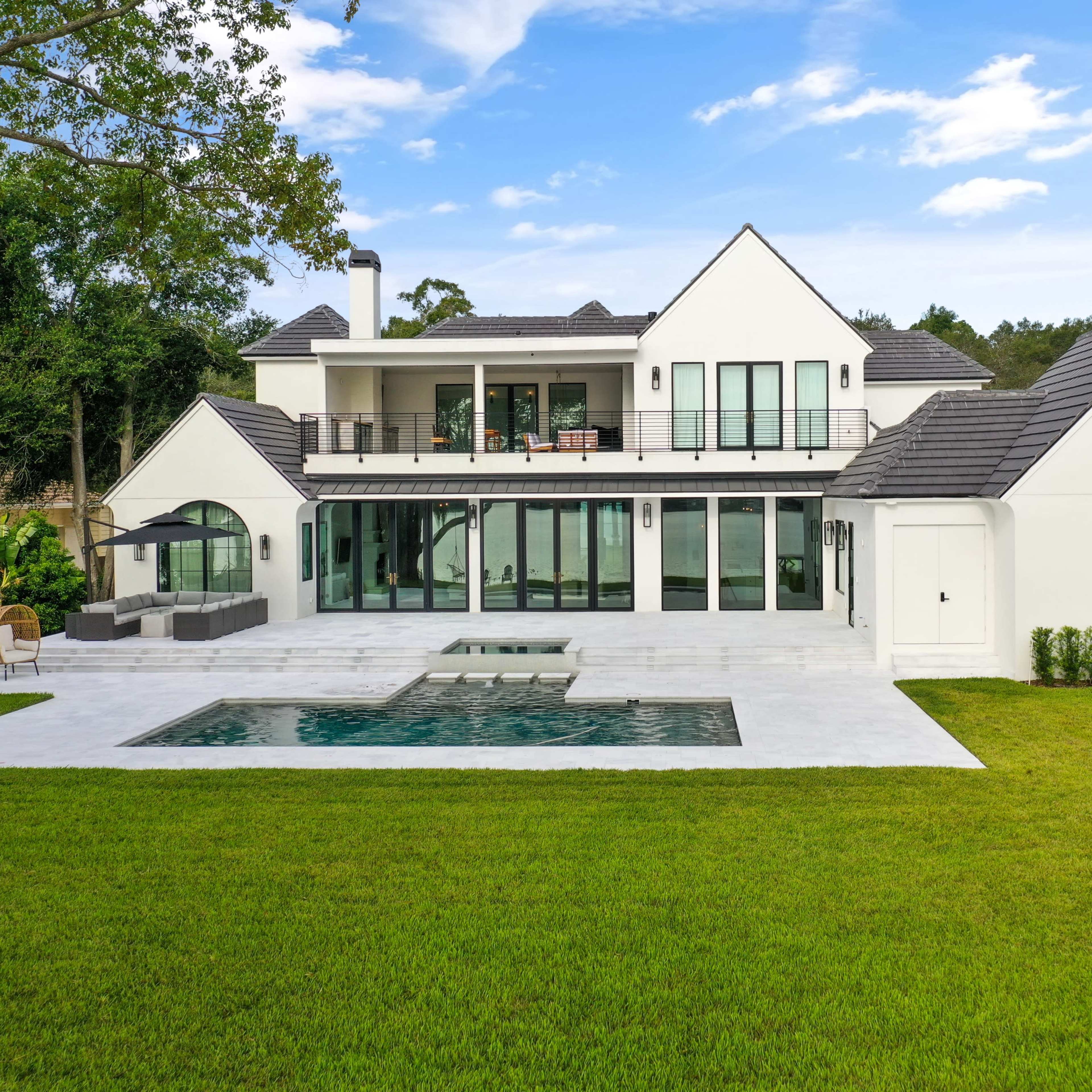 A modern two-story house with large windows and a swimming pool is set on a grassy lawn surrounded by trees.
