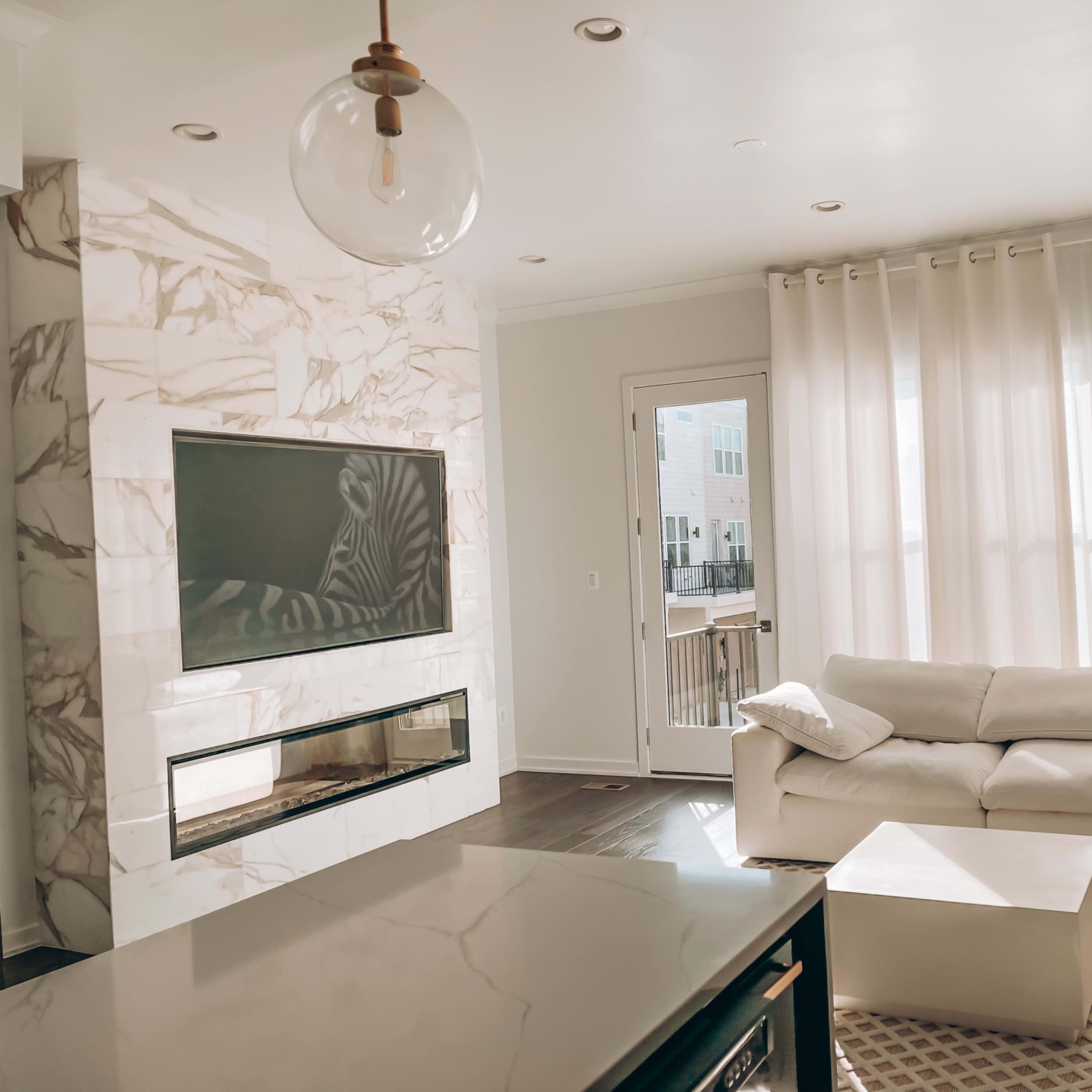 A modern living room features a marble accent wall with a flat-screen TV, large windows draped with sheer curtains, and a white sectional sofa.