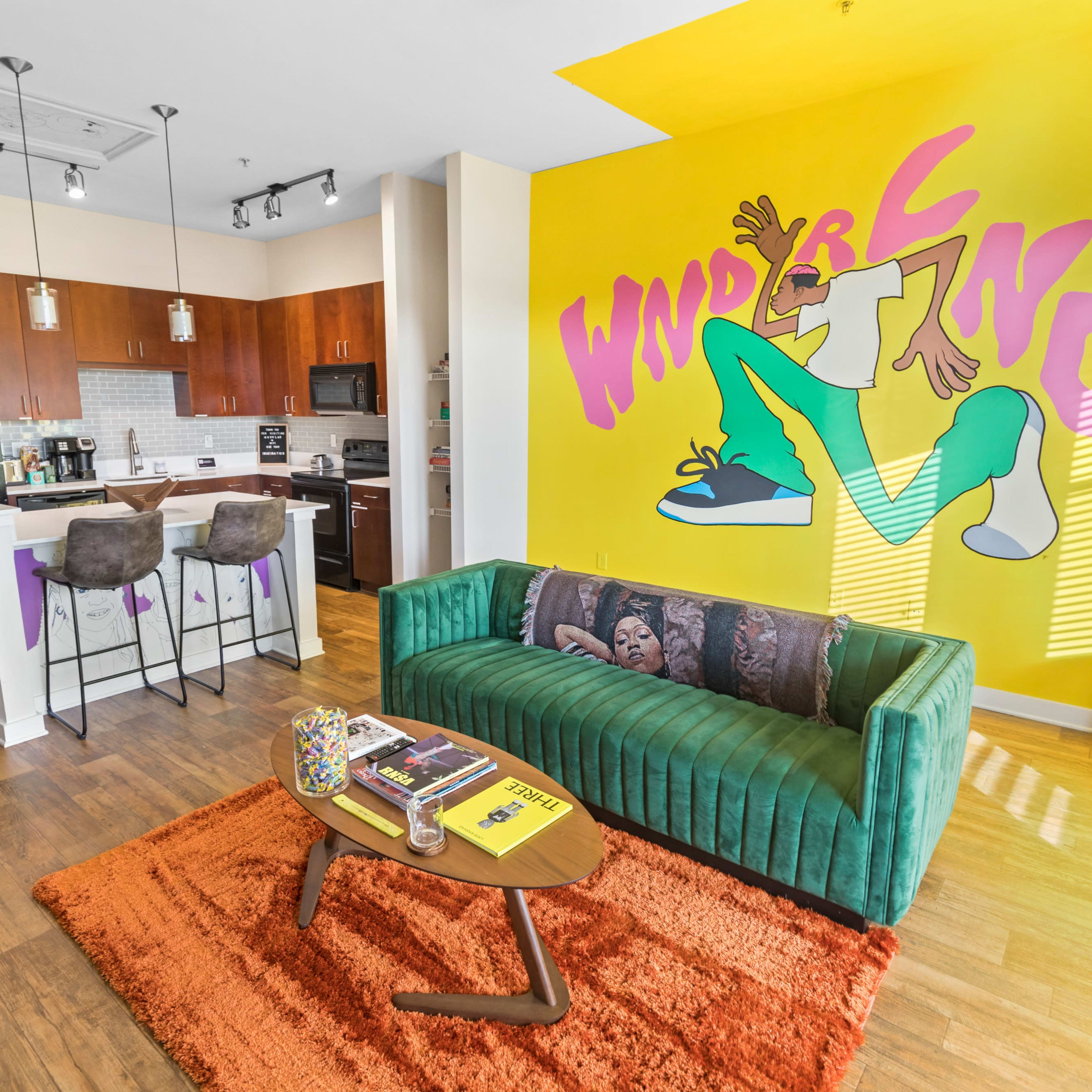 A modern apartment living area with a green couch, a wooden coffee table, and a kitchen space featuring dark cabinets and stainless steel appliances, highlighted by a bright yellow wall with cartoon-style lettering.