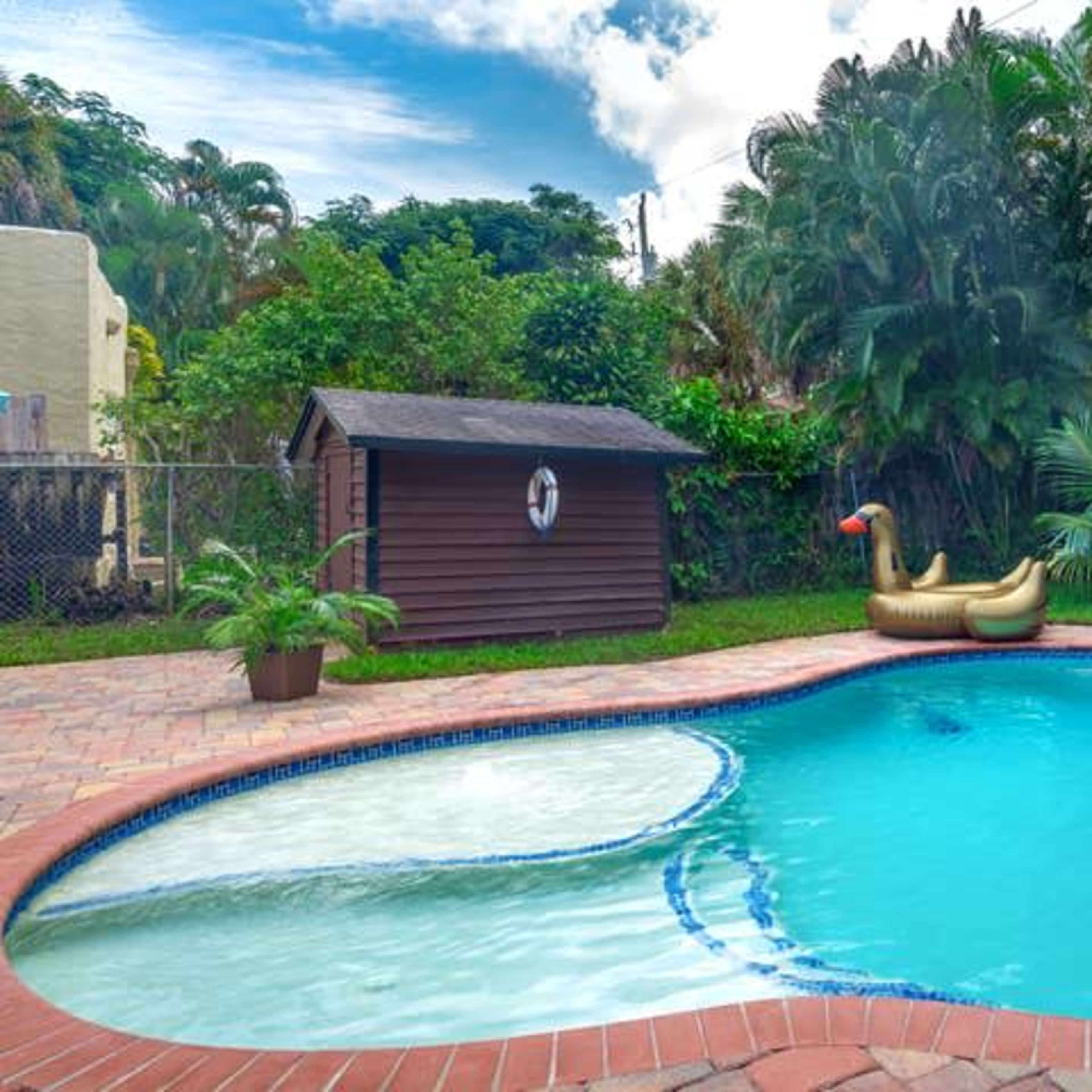 This scene features a swimming pool surrounded by lush greenery, with a large inflatable swan float and two blue lounge chairs nearby.