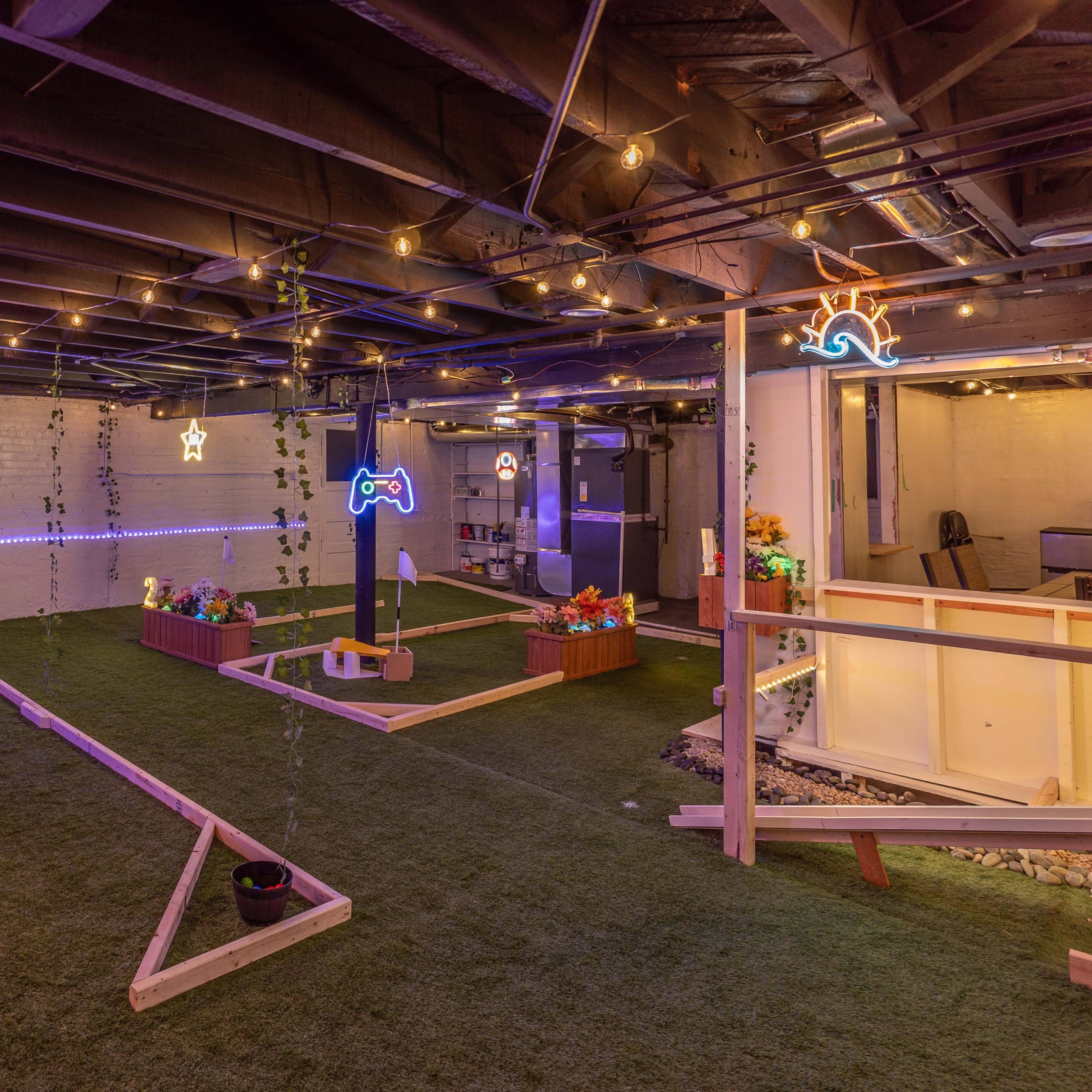 A basement space decorated with colorful neon lights, artificial grass, and wooden planters filled with flowers.