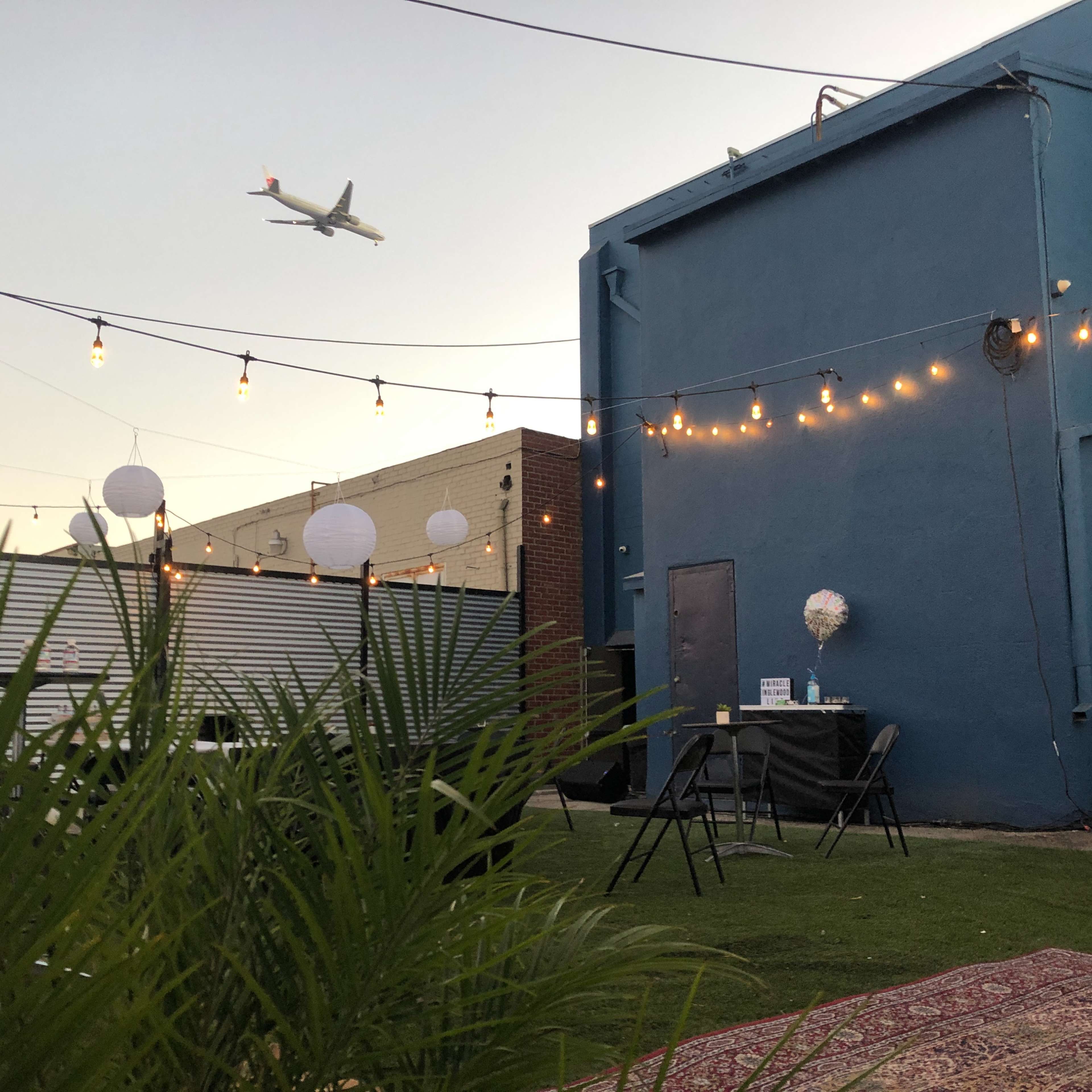 A decorated outdoor space with string lights, a table, and greenery, with an airplane flying overhead.