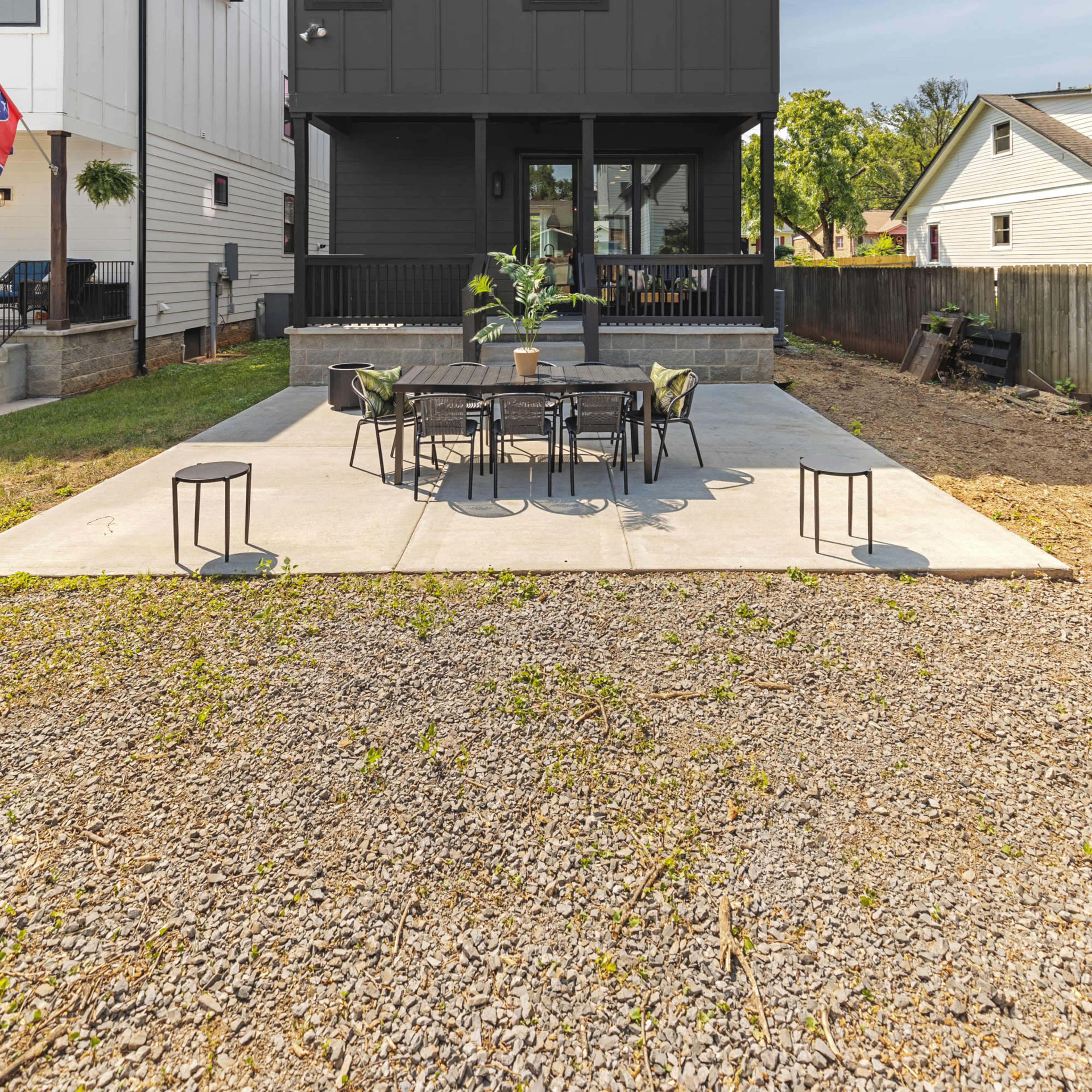 A modern two-story black house is situated behind a gravel yard with a concrete patio and outdoor dining set.