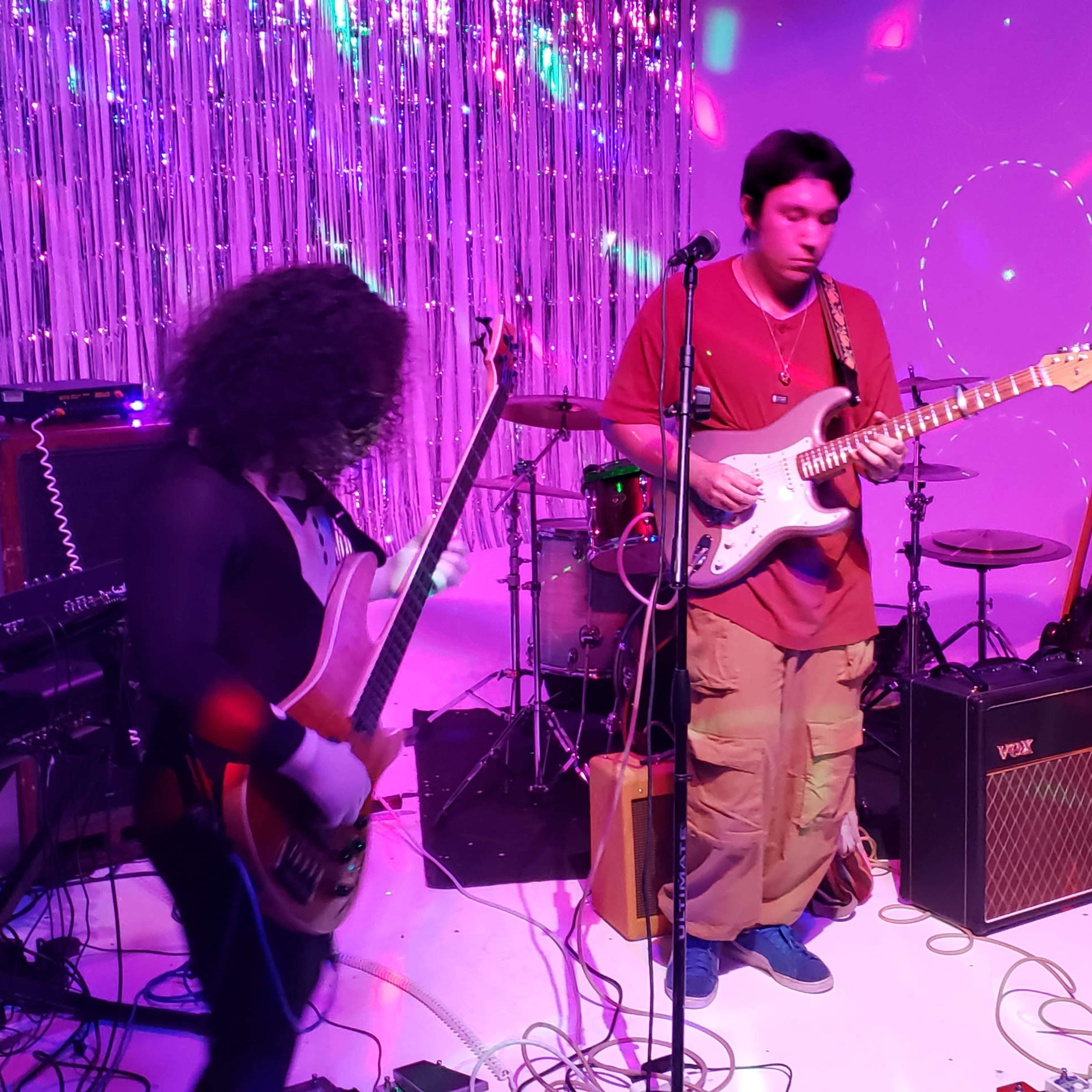 A music band performs on stage with various instruments in front of colorful lights and a shimmering backdrop.