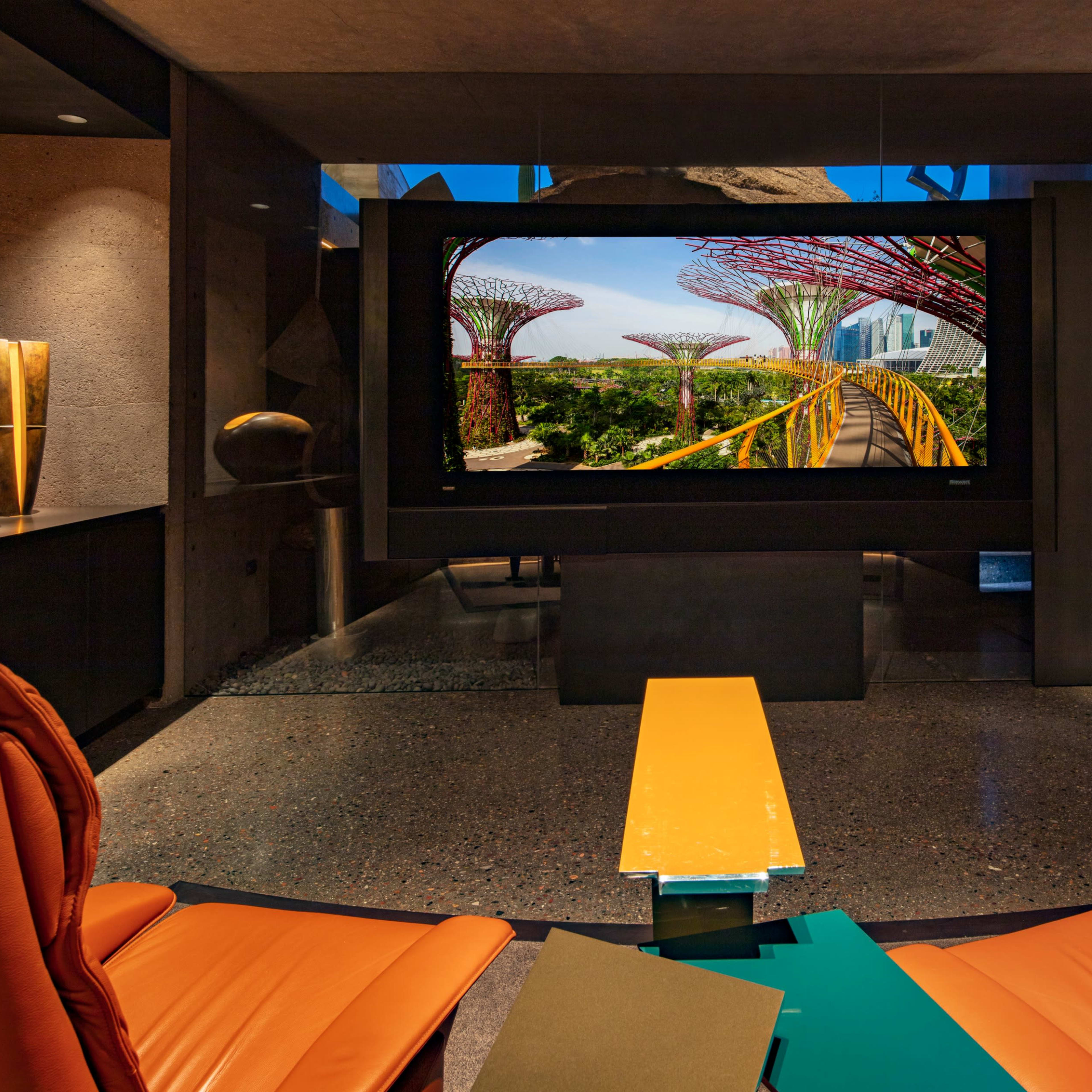 A modern interior featuring two orange chairs facing a large screen displaying a vibrant garden scene with tall structures in the background.