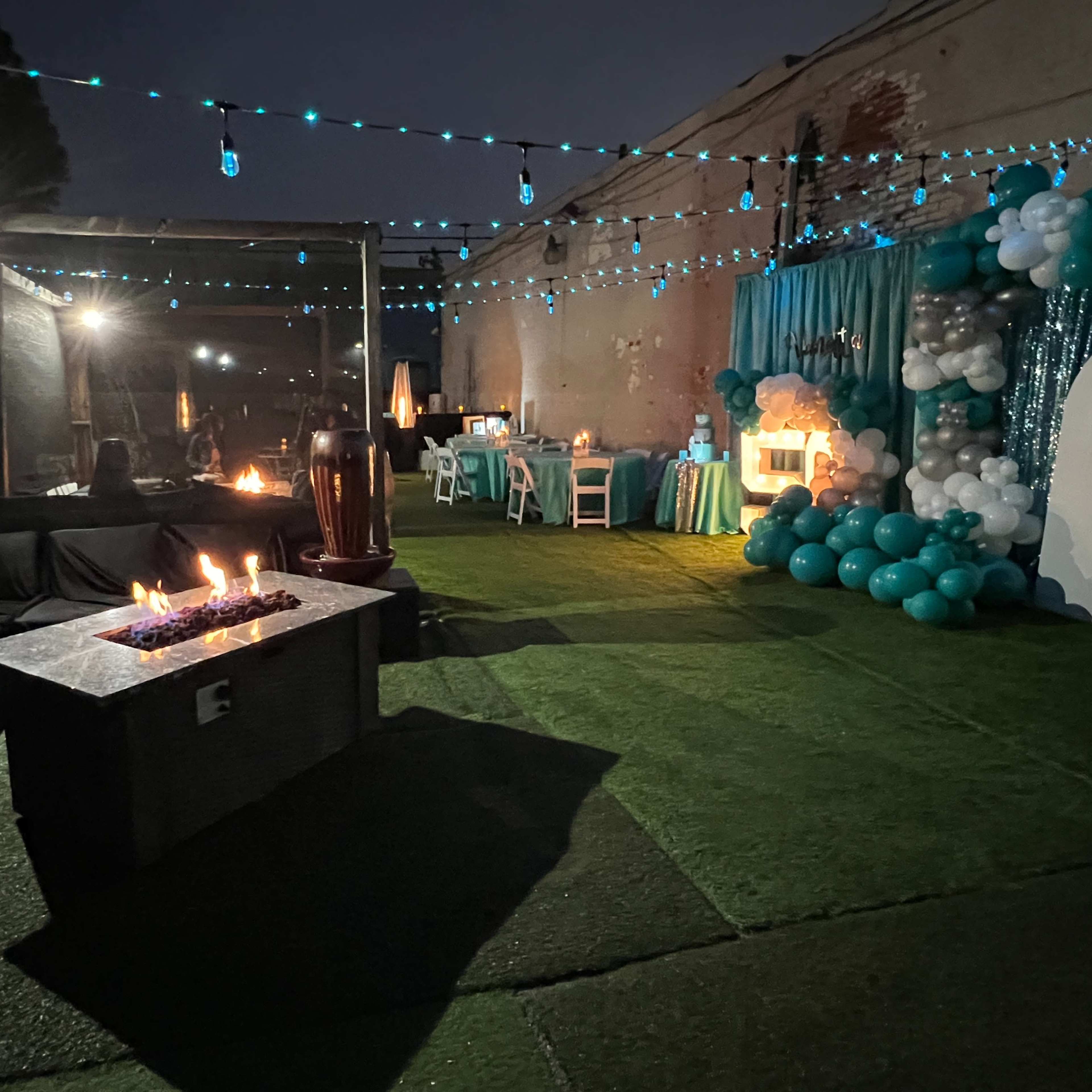 An outdoor party setup featuring a fire pit, a seating area with a dark sofa, tables decorated with teal balloons, and ambient lighting.
