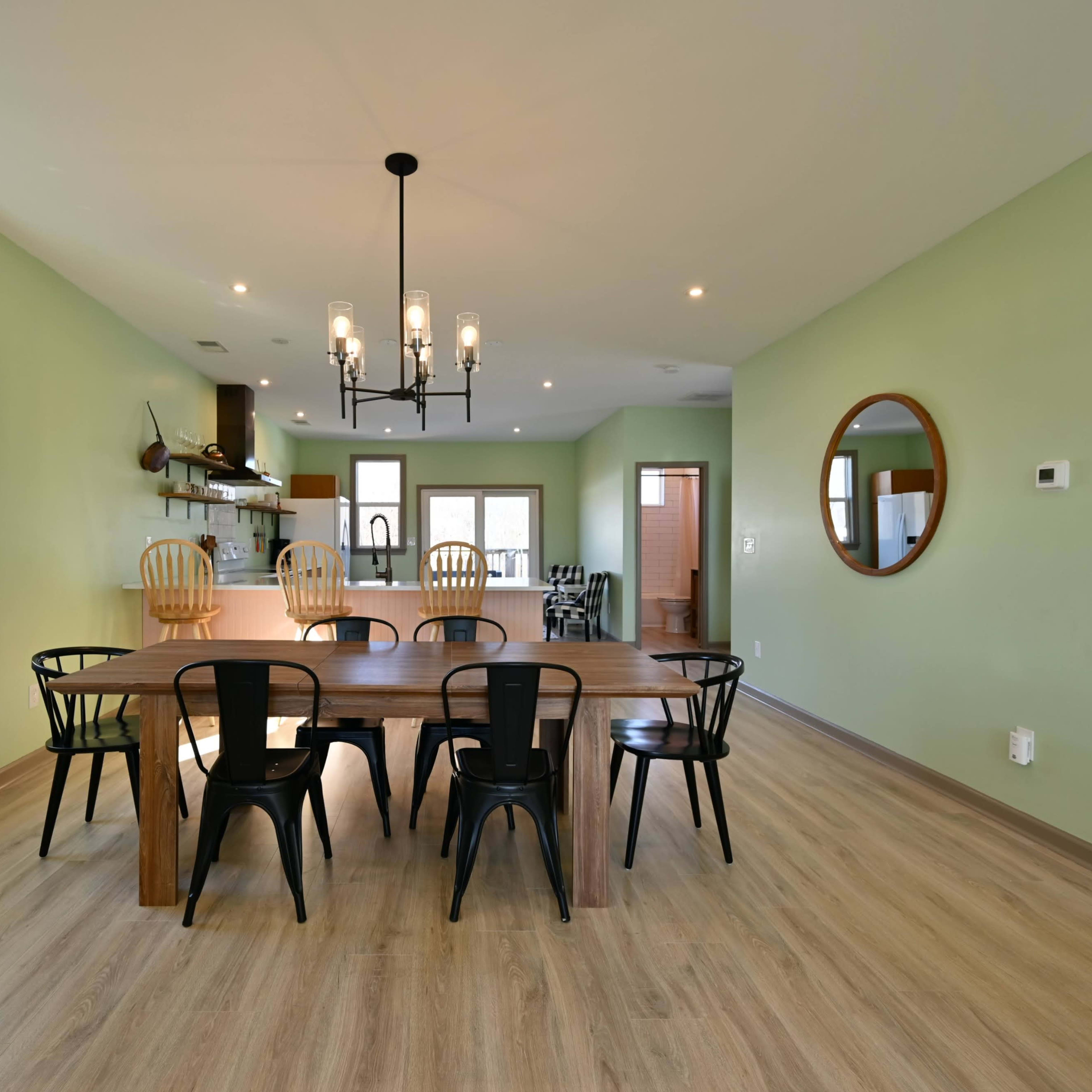 A spacious dining area features a long wooden table surrounded by black and wooden chairs, with green walls and modern lighting fixtures above.