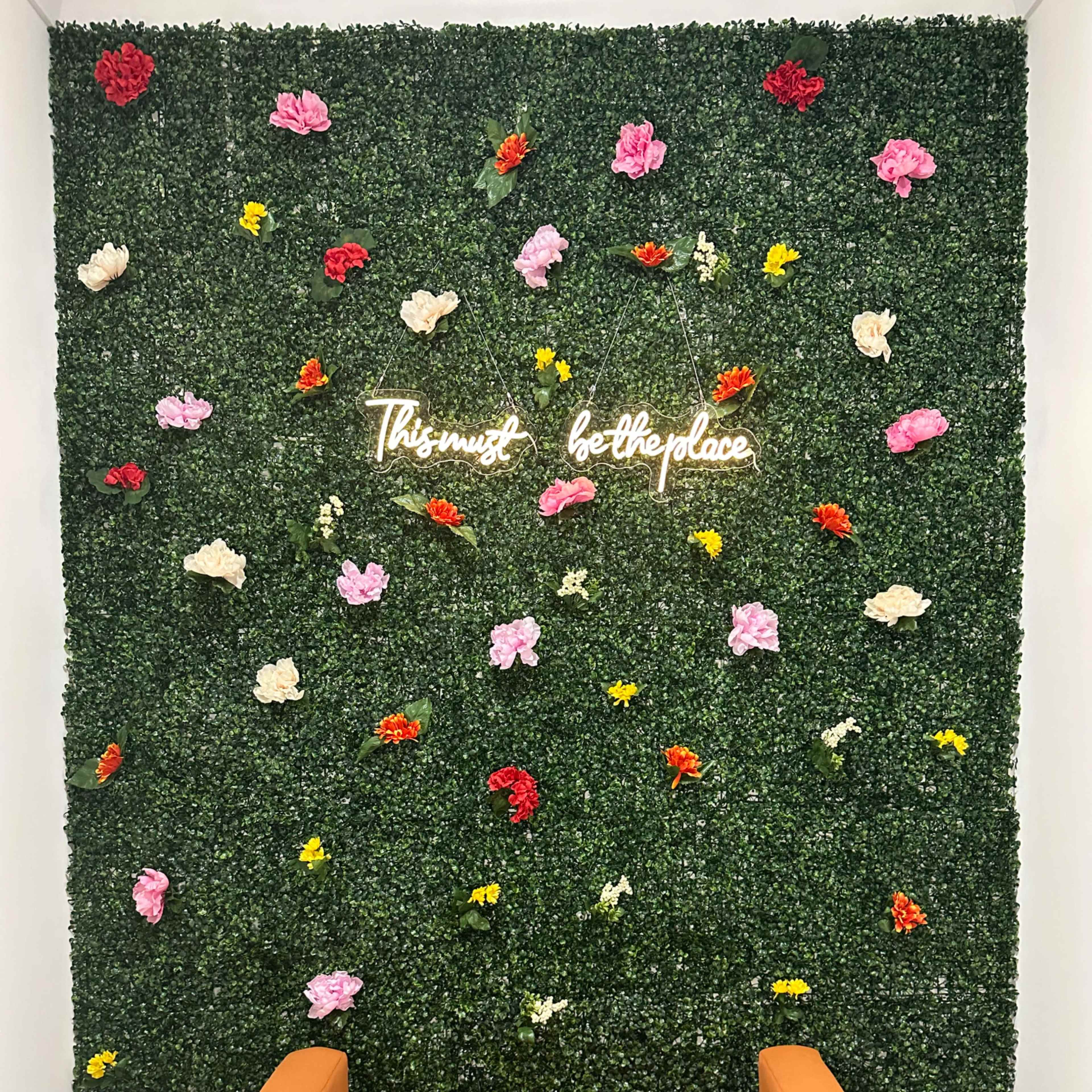 An orange couch in front of a green wall adorned with artificial flowers and a neon sign that reads "This must be the place."