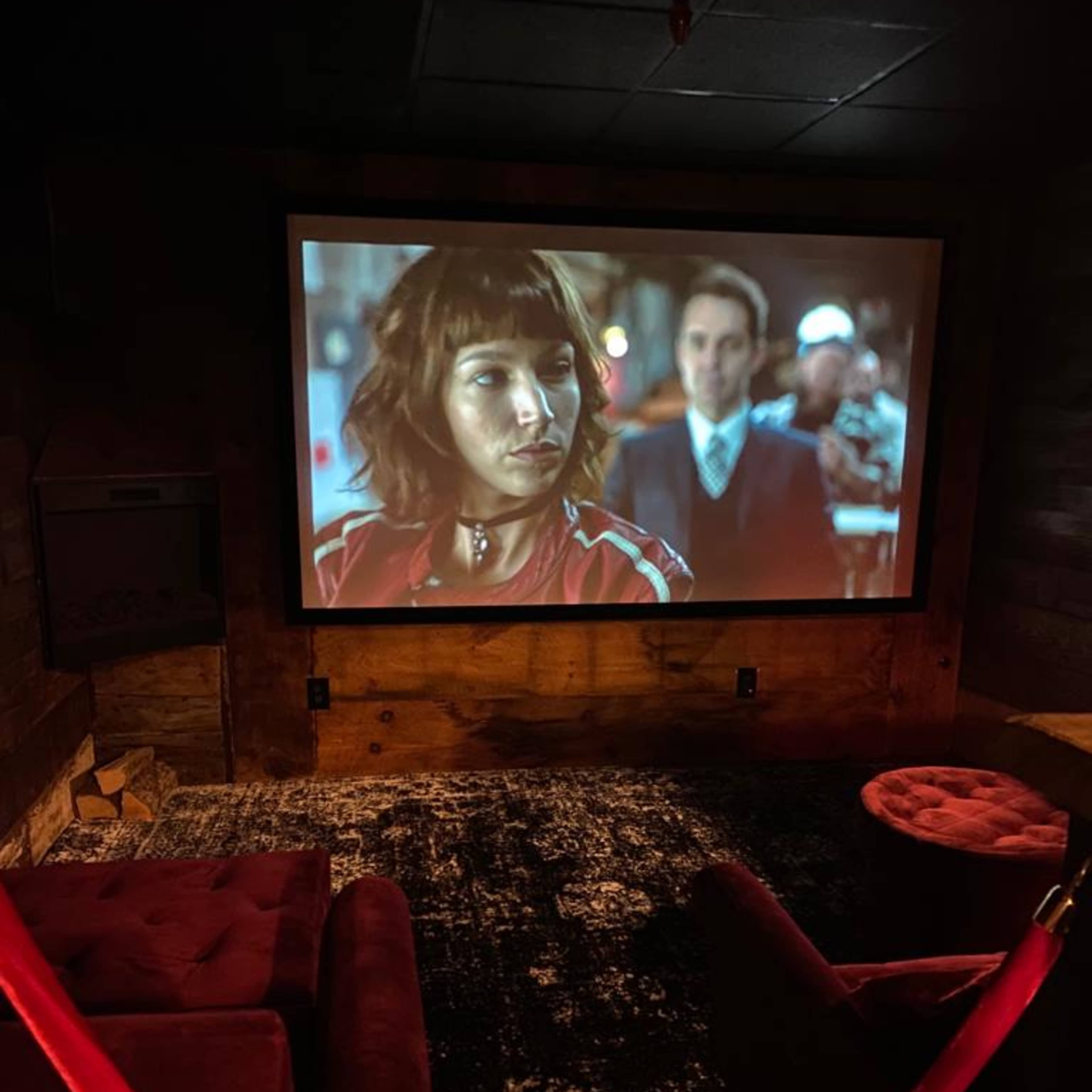 A home theater setup features a large screen displaying a movie scene, with red velvet seating and a red rope barrier in front.