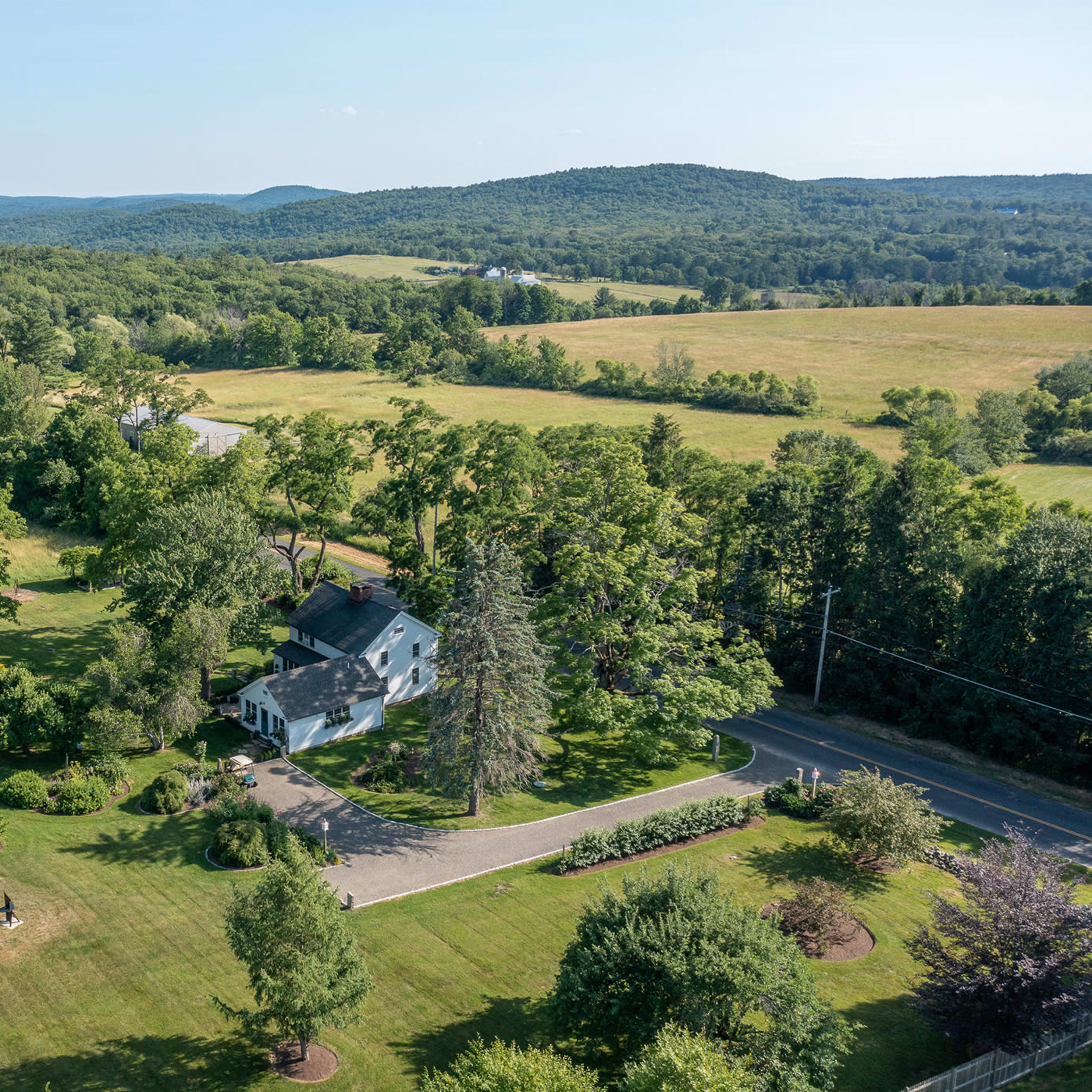 An aerial view shows a house surrounded by lush trees and fields, with a winding road visible nearby.