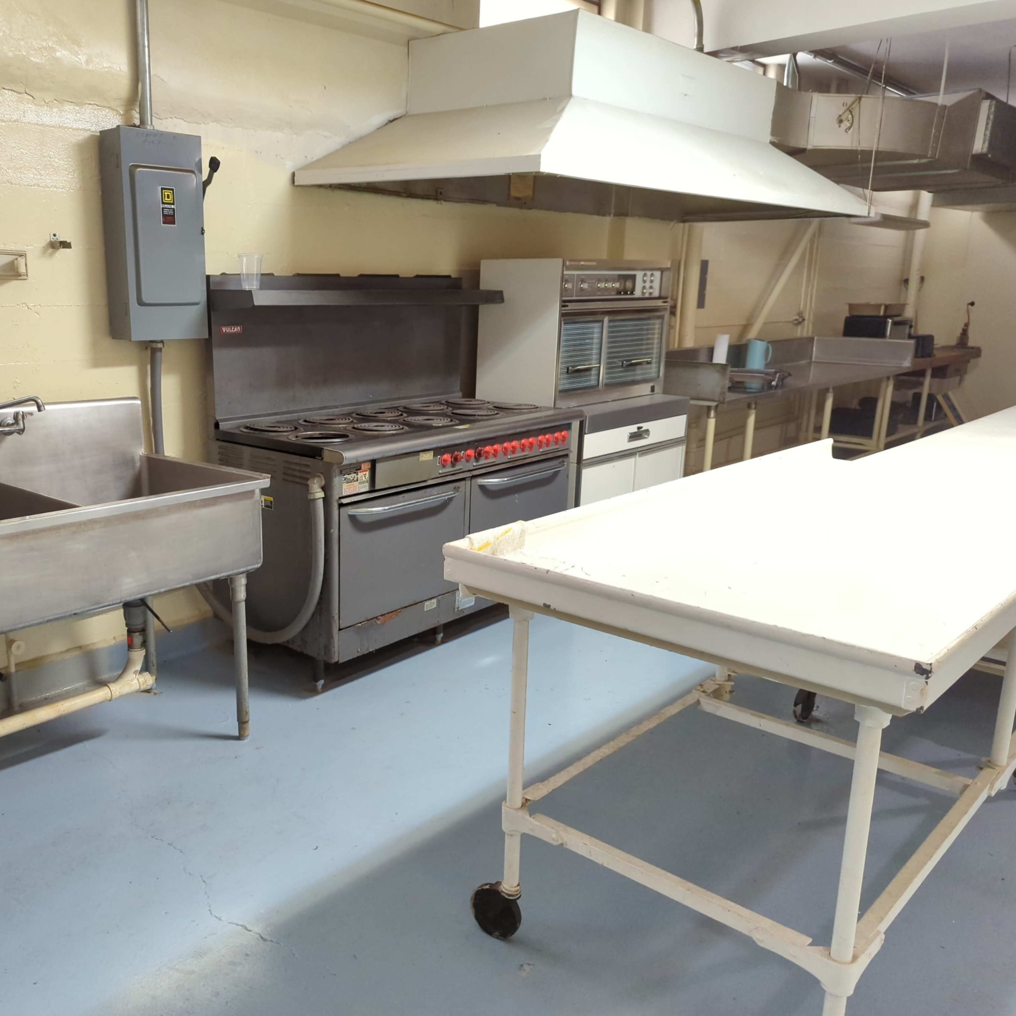 A commercial kitchen equipped with a stainless steel sink, a gas range with multiple burners, a metal work table, and ventilation hoods.