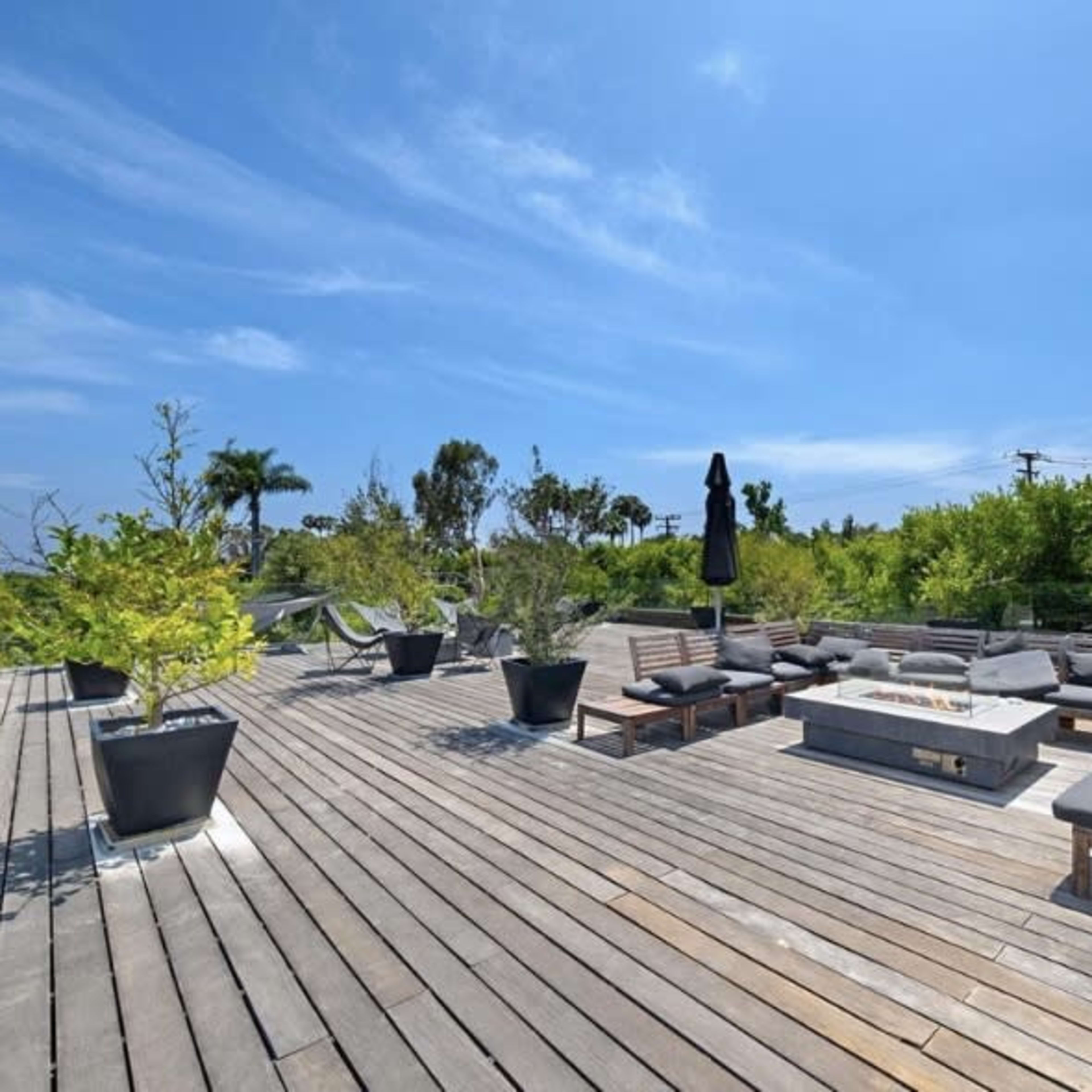 A spacious wooden deck features seating areas with cushions, potted plants, and a central fire pit under a clear blue sky.