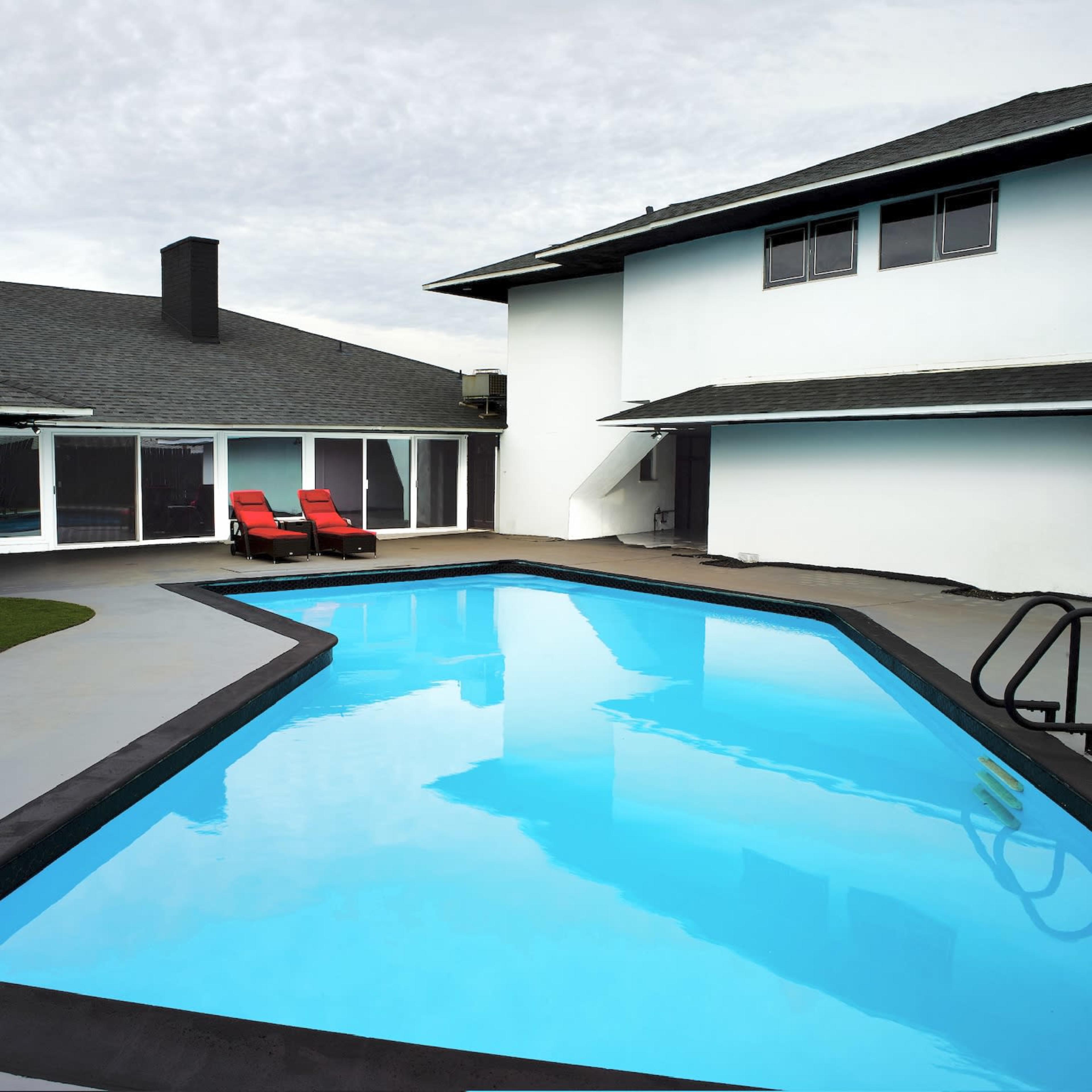 A modern backyard featuring a swimming pool bordered by a concrete patio and two red lounge chairs.