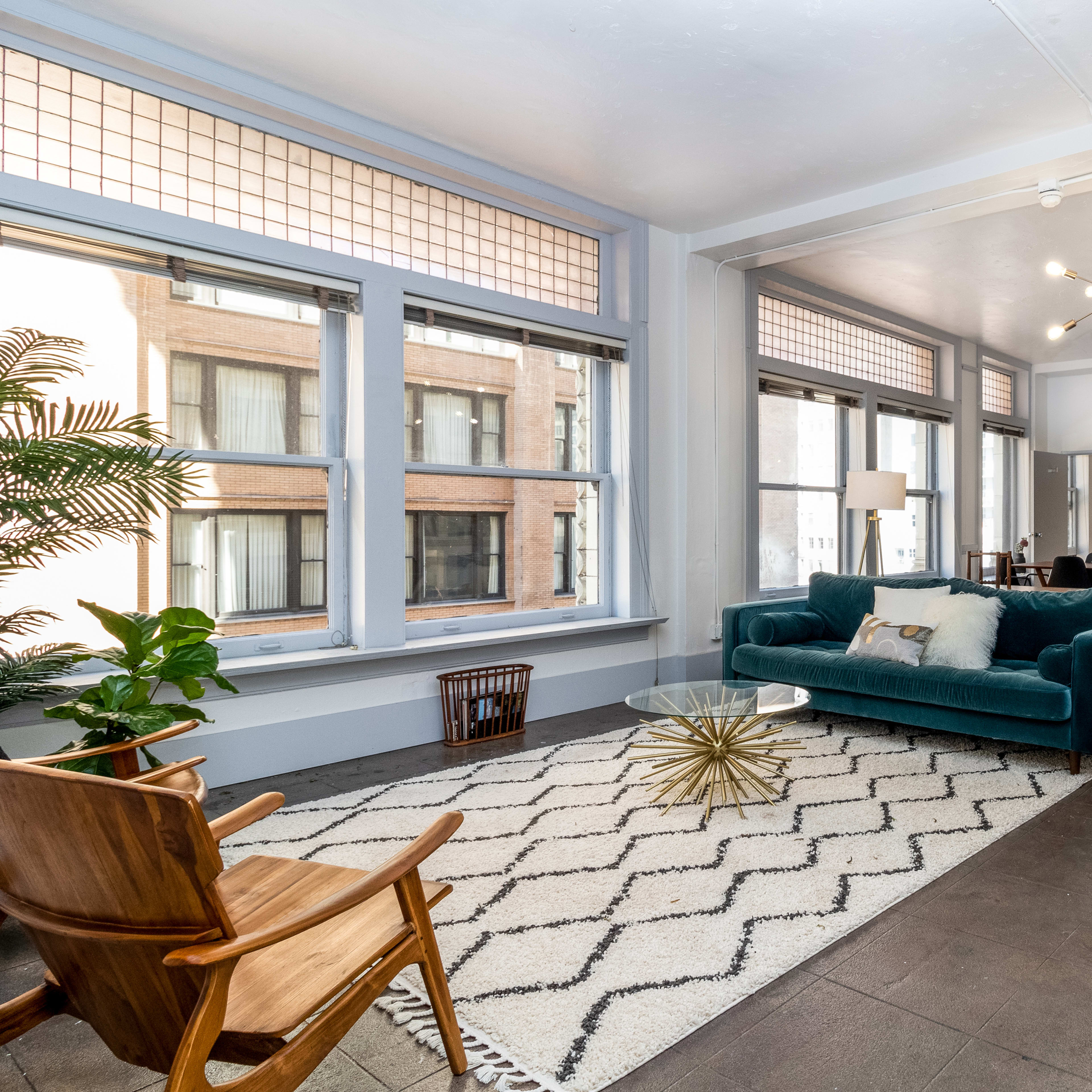 A bright, modern living space features large windows, a teal couch, a geometric rug, and a dining area in the background.